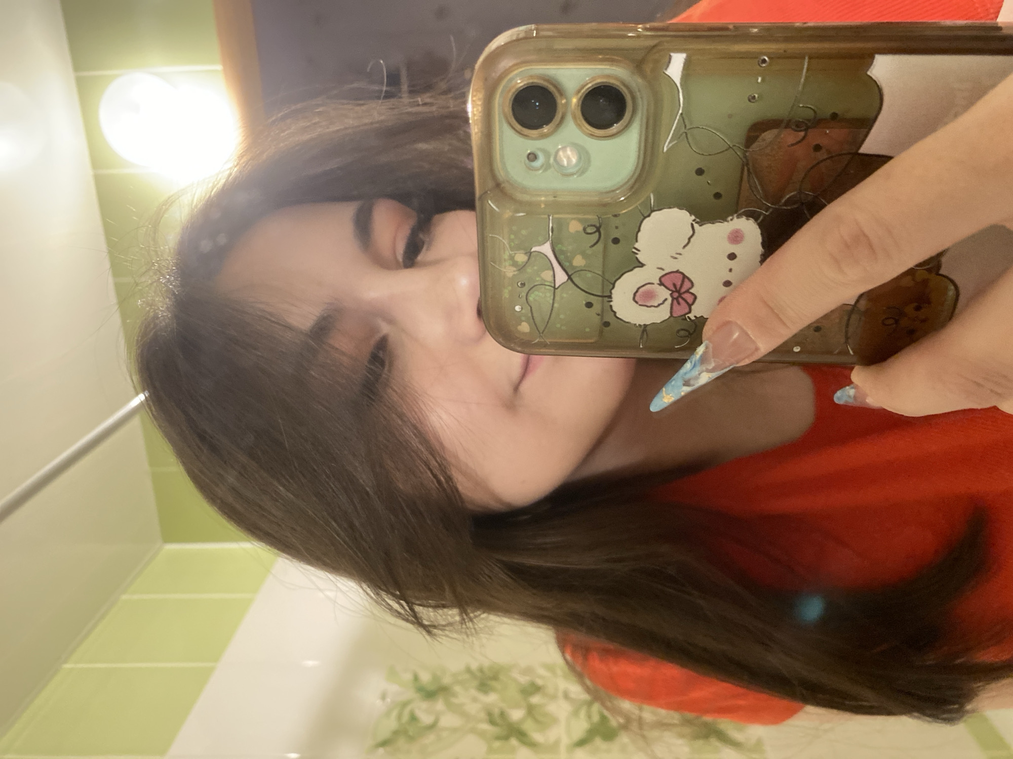 A person is taking a selfie in a bathroom. The individual is wearing a red shirt and has long, dark brown hair that falls over their face. Their fingernails are long with a blue and white design. They are holding a phone with a light green case decorated with a cartoon character, possibly Hello Kitty, in front of their face. The background includes a light fixture and green and white tiles. The location appears to be indoors and is likely a private residence. The city of Chaikovsky, Russia, cannot be determined from the image alone.