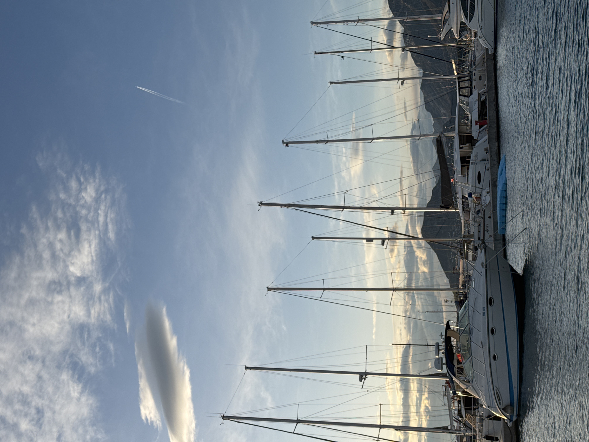 The image shows a marina in Bar, Montenegro, featuring a row of sailboats docked along a pier. Their masts extend upwards into a sky with scattered clouds, including a prominent, vertical cloud formation. In the distance, mountains are visible, partially obscured by haze or clouds. A contrail from an airplane cuts across the upper left of the sky. The water in the marina reflects the sky and boats.