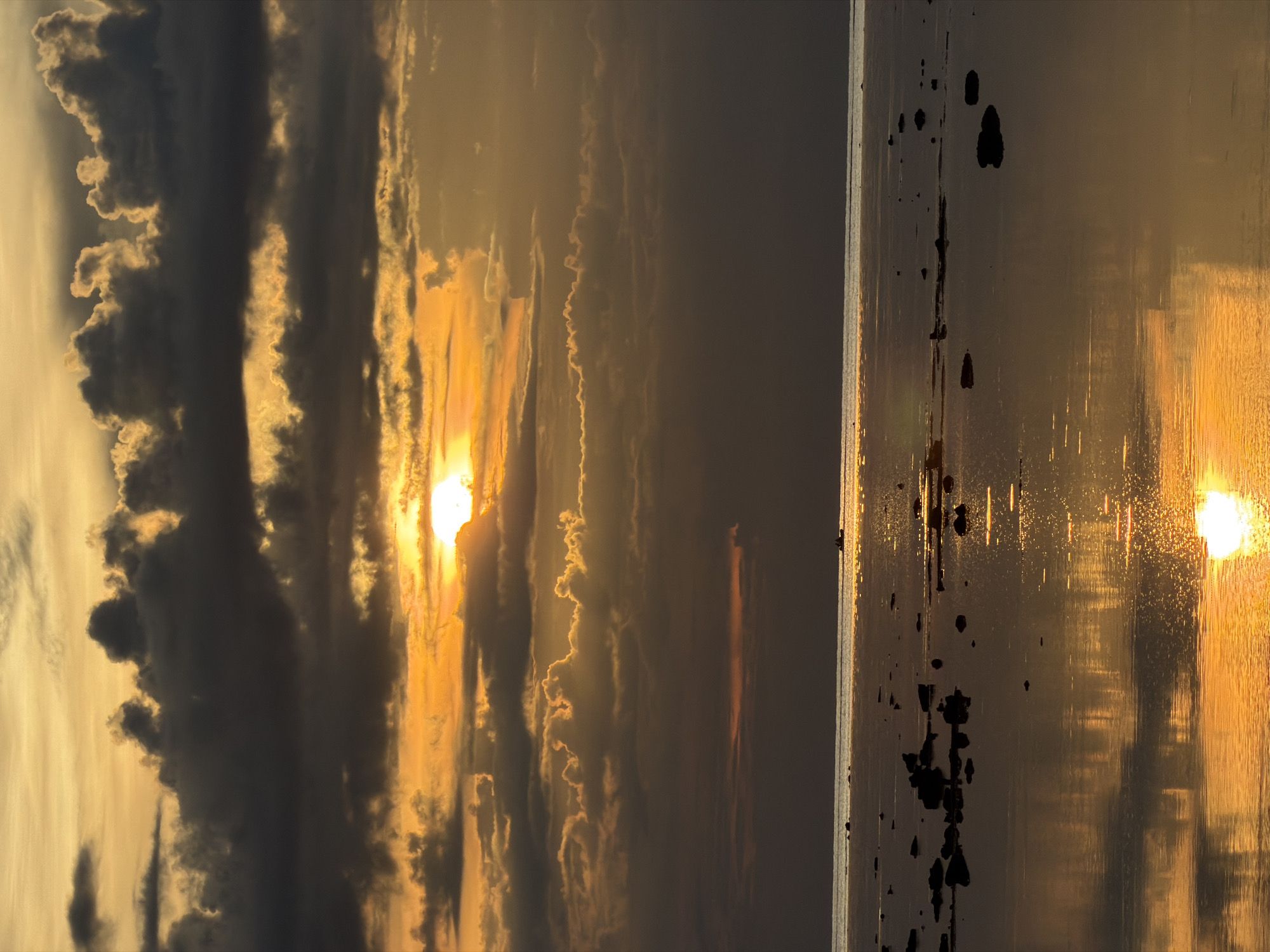 The image depicts a breathtaking sunset over a body of water, likely the sea, as suggested by the reflection of the sun and scattered dark objects resembling rocks or debris in the water. The scene is divided vertically. The left side showcases a dramatic sky filled with a mix of dark, heavy clouds and lighter, wispy clouds illuminated by the setting sun. The sun itself is a bright, golden orb peeking through the clouds, casting warm orange and yellow hues across the sky and reflecting onto the water. The right side of the image is dominated by the shimmering water surface, reflecting the vibrant colors of the sunset. Dark, irregular shapes are scattered across the water, adding a sense of texture and depth. The overall mood is serene and awe-inspiring, characteristic of a beautiful sunset. The time of day is clearly sunset, with the low-lying sun creating warm, golden light. The weather appears partly cloudy, contributing to the dramatic visual effect. The location context of Baan Plai Laem, Thailand, suggests a coastal or island setting. There is no visible text in the image.