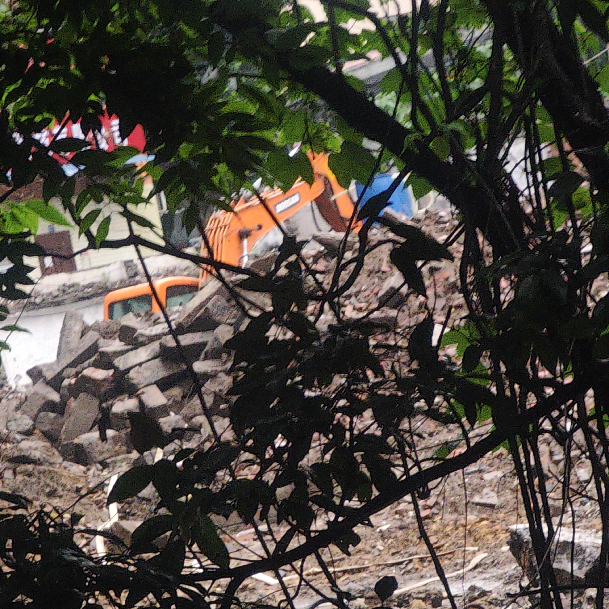 The image provides an obscured view of what appears to be a demolition or construction site, heavily framed and partially hidden by dense green foliage and dark tree branches in the foreground.
In the mid-ground, two orange heavy machinery vehicles are visible among extensive piles of gray and brown rubble, concrete, and earth. One prominent orange excavator is seen with the brand name "DOOSAN" clearly written in white letters on its arm. Its bucket is positioned towards the debris. To the left, the cab of another orange vehicle, possibly a second excavator or a large truck, is partially visible, appearing to be situated within or on top of a separate pile of rubble.
The ground around the debris appears uneven and somewhat damp in areas. In the background, beyond the immediate work area, glimpses of structures can be seen, including elements with red and white striped patterns, possibly parts of buildings or awnings, and a blue tarp or object. The scene is illuminated by daylight, but the heavy foreground vegetation creates a filtered and somewhat dim light. No people are visible. The overall impression is of an active industrial or cleanup site, viewed from a distance through natural cover.