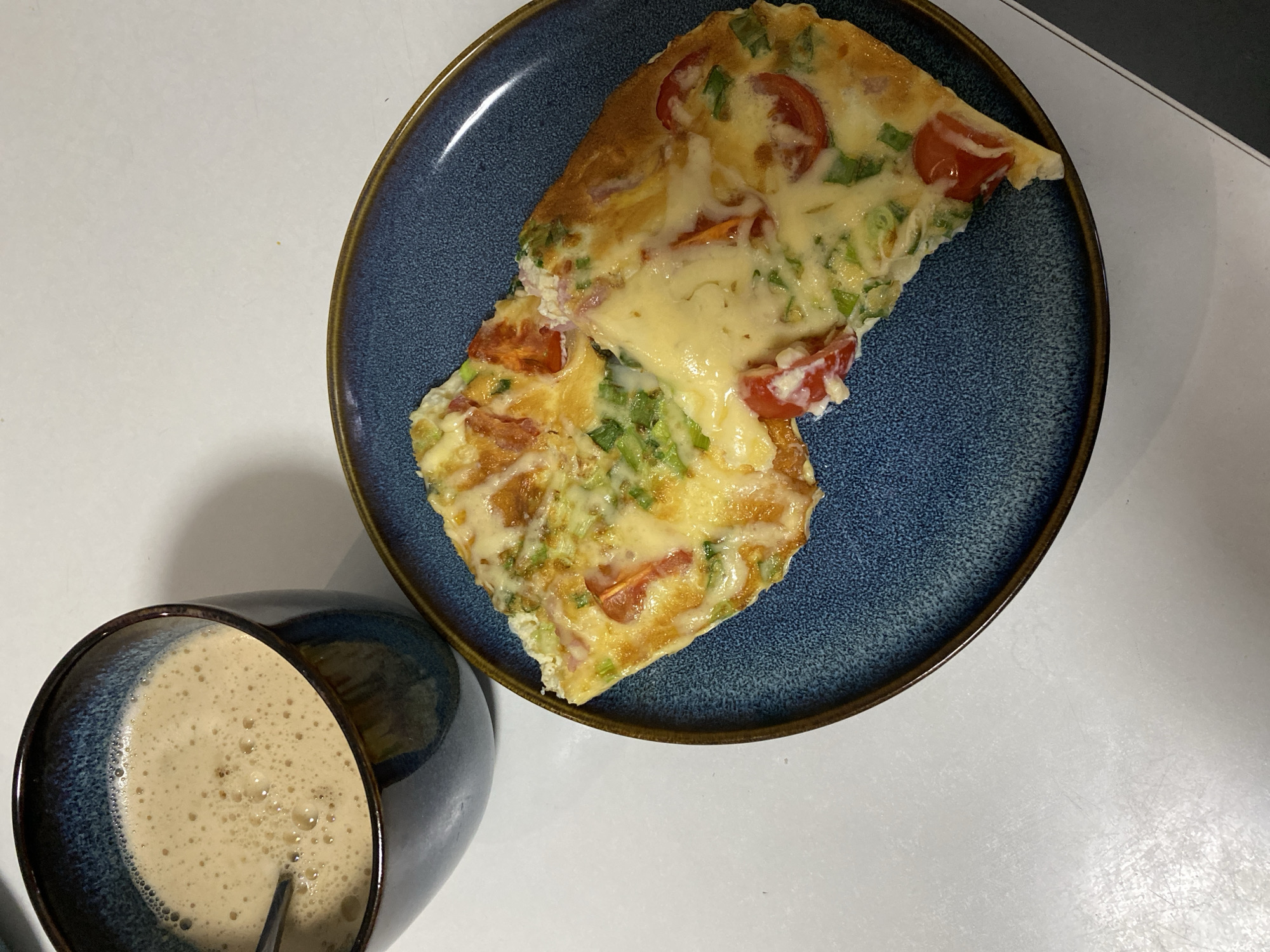 A top-down shot shows a blue ceramic plate with a slice of frittata topped with melted cheese, tomatoes, and green onions. Beside the plate is a dark blue ceramic mug filled with a frothy beverage, possibly coffee or latte. The plate and mug are on a white surface. The image does not depict a specific location in Noviy, Russia, or any people or actions.