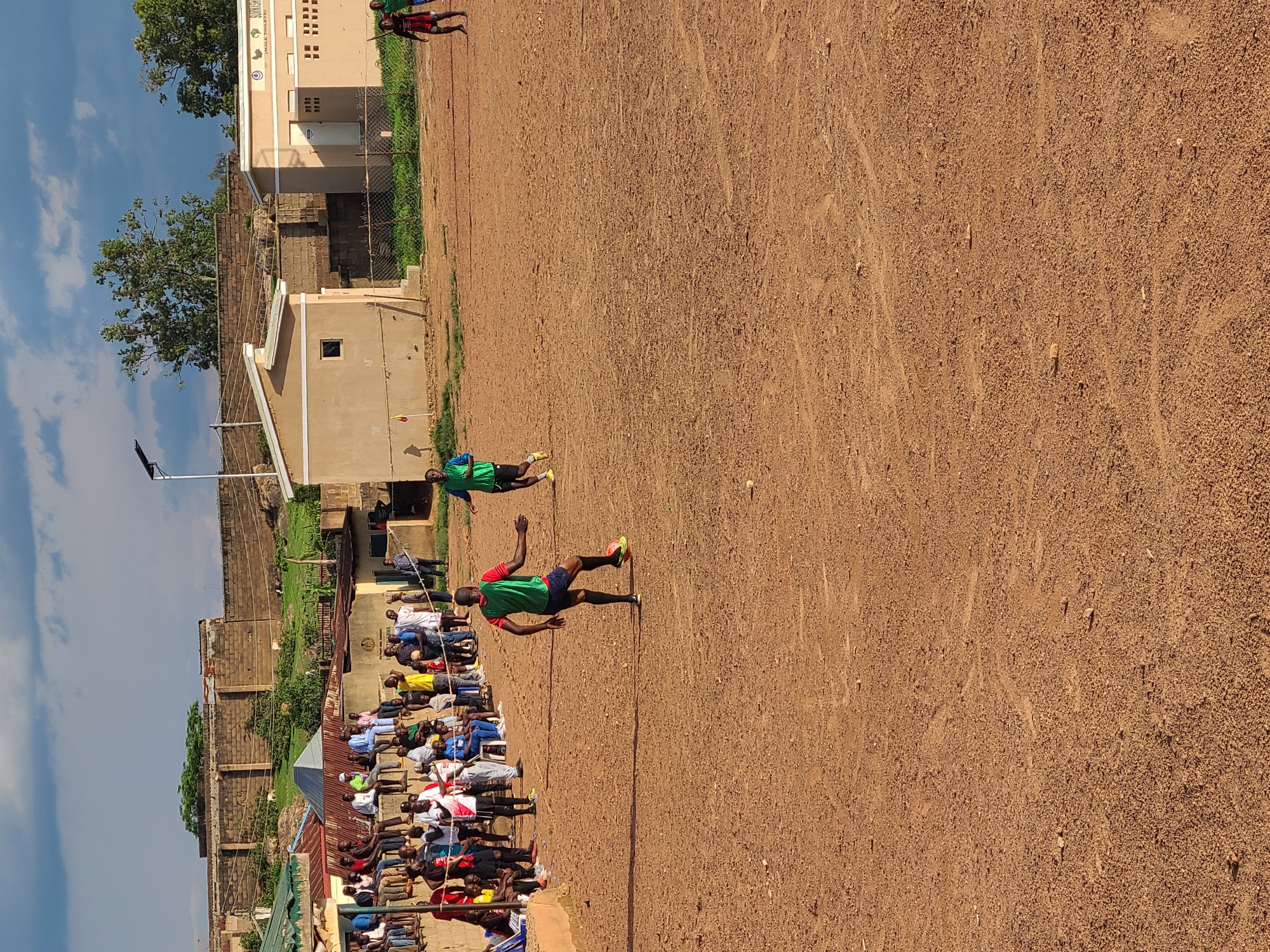 This image captures an outdoor scene on a dry, reddish-brown dirt field, consistent with an informal soccer pitch, at an unknown location within an unknown city and country. The sky is partially cloudy, featuring patches of blue interspersed with darker clouds, and the lighting suggests late afternoon or early morning, casting distinct shadows.
In the foreground, slightly left of center, a male individual is actively dribbling a yellow soccer ball with his right foot. He wears a green and red sleeveless vest over a dark shirt, dark shorts, and bright yellow-green athletic shoes. Another male individual in a bright green sleeveless vest, dark shorts, and yellow-green athletic shoes is running in the mid-ground, right of center. A third male individual, wearing a red and black outfit, is visible running near the far-right edge of the frame. All three individuals appear to be of Black ethnicity.
Along the left side of the field, approximately 50 to 70 spectators, predominantly male, are seated or standing on a raised concrete barrier or wall, observing the activity on the field. They are casually dressed in various shirts, t-shirts, and pants. Behind them, a series of buildings with corrugated metal roofs are visible, interspersed with some greenery. Further back, a long, tall wall constructed of unrendered concrete blocks or bricks forms a significant horizontal barrier across the mid-ground.
On the right side of the image, two single-story, tan-colored buildings with flat roofs are situated behind a chain-link fence. The building on the right features a white door and ventilation blocks, and a sign on its facade indicates "SURKHSHA" and "(MALE TOILETS)". The building to its left has a small, single window. A tall pole equipped with a solar panel and an LED light fixture stands behind the left tan building, suggesting a street light or area illumination. A yellow and red corner flag is visible on the field near the left tan building. The ground of the field shows traces of tire tracks or worn paths.