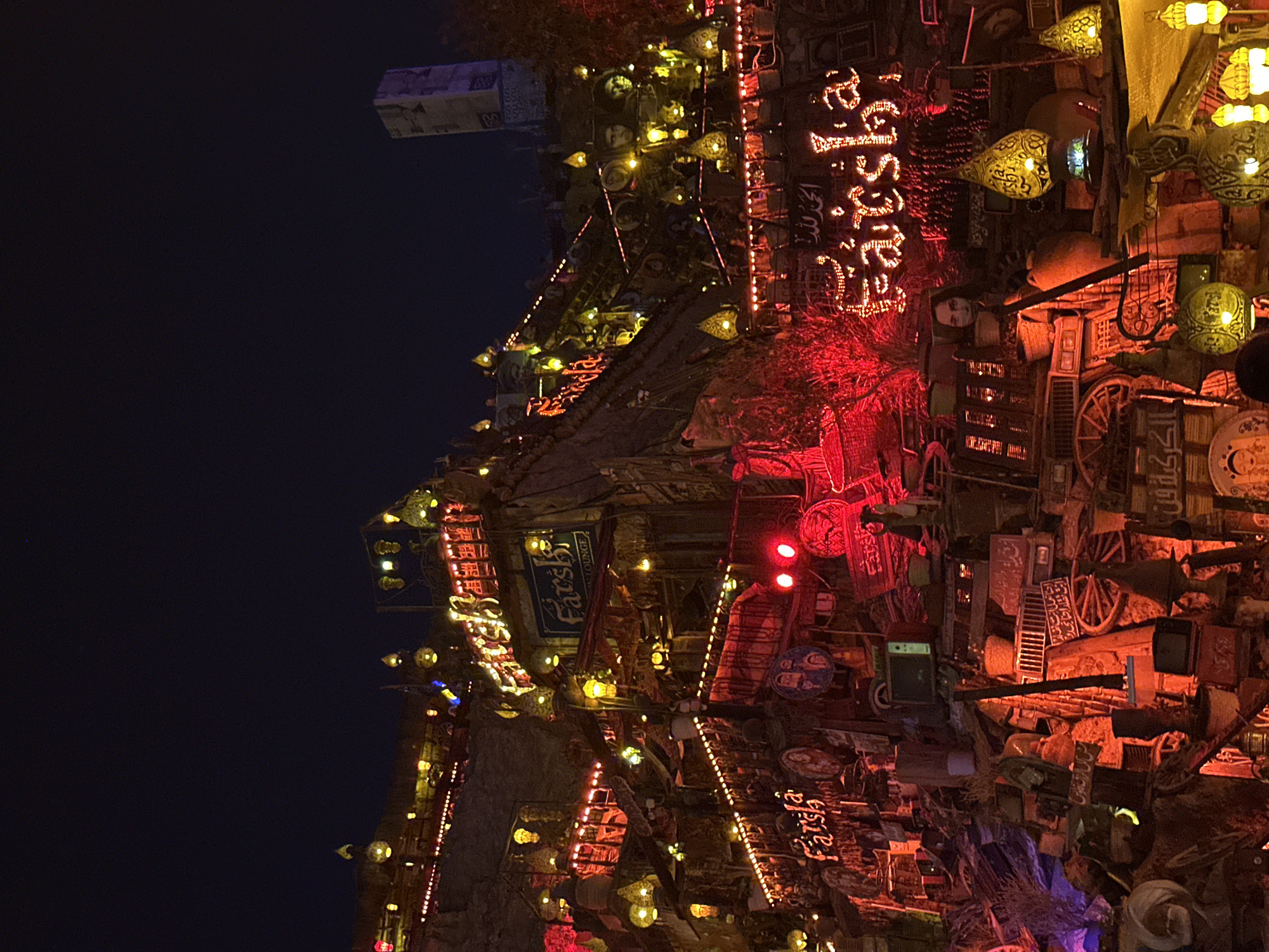 This media file depicts an elaborately decorated outdoor cafe or lounge at night, likely in Sharm El Sheikh, Egypt. The scene is illuminated by abundant artificial lighting, primarily warm yellow, orange, and red hues from various lanterns, string lights, and neon signs, creating a cozy and exotic ambiance against the dark night sky.
The structures appear multi-layered, possibly built into a hillside, and are densely adorned with an eclectic collection of objects. These decorations include traditional pottery, ornate metal lanterns, old wooden wheels, framed artwork, discarded electronics, and other salvaged items, contributing to a unique, bohemian aesthetic. Red foliage or fabric elements are integrated into the decor, adding texture and warmth. A prominent feature is a mannequin head peering from behind some decorations.
Several signs are visible, including the name "Farsha" in both English and prominent red Arabic script. Other legible Arabic texts include "Farsha LOUNGE," "مش كل الرزق فلوس" (Not all blessings are money), "الكركية فن" (Al-Karkaya is an art/craft), and "الحمد لله" (Praise be to God).
The scene suggests a vibrant gathering place for socializing and relaxation, designed to offer a unique cultural experience in a visually stimulating environment. The warm lighting and rich, detailed decor evoke feelings of charm and tranquility.