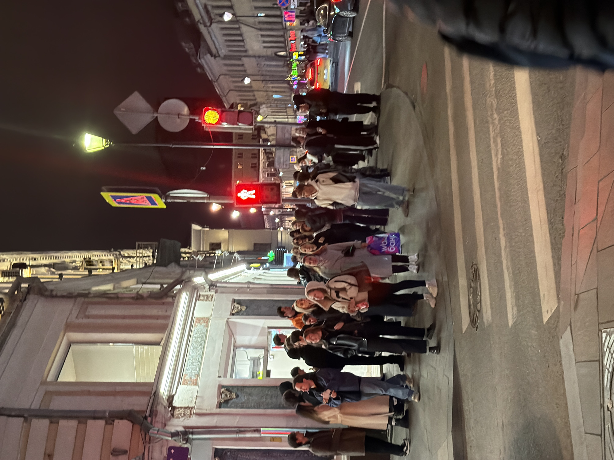 A crowd of pedestrians is waiting at a crosswalk in Moscow, Russia. The traffic light displays a red light, indicating pedestrians should not cross. The photo was taken at night; a street light and the lights of the buildings illuminate the scene. The surrounding buildings, street signs, and crosswalk stripes are visible. The people are standing on the sidewalk at the intersection, waiting for the light to change. Most of the pedestrians are facing the same direction, ready to cross the street.