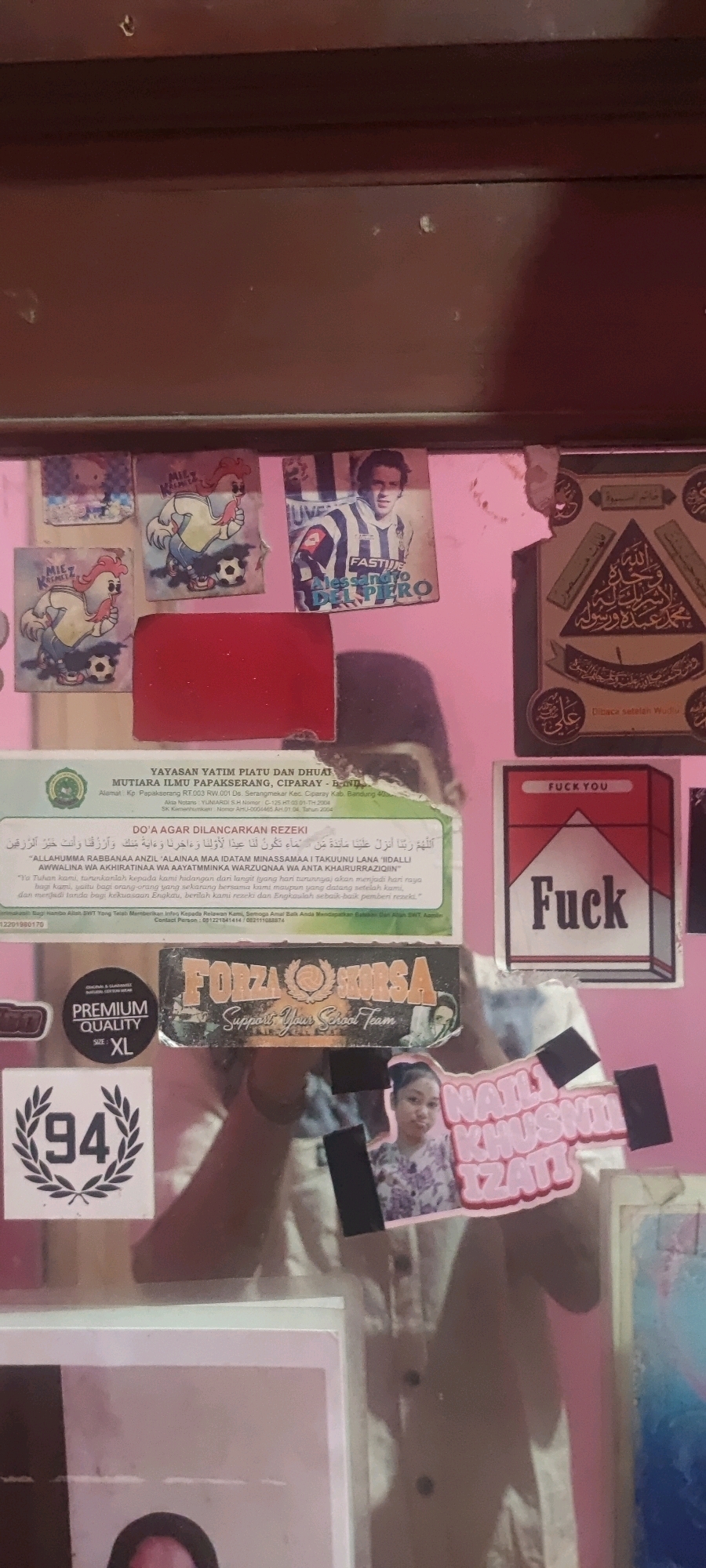 The image shows a close-up of a wall decorated with various stickers and a prayer banner. The wall is painted pink and appears to be indoors, possibly in a bedroom or living area. In the top left quadrant, there are several stickers, including two featuring cartoon chickens playing soccer, one with a Hello Kitty character, and a poster of the soccer player Alessandro Del Piero. Below these stickers, a red rectangular object is affixed to the wall.
To the right of the Del Piero poster is an Islamic calligraphy banner with intricate designs. Further down, a sticker resembling a cigarette pack with the words "Fuck You" is visible. In the bottom left quadrant, a sticker with the number "94" inside a laurel wreath is present. Another sticker below it reads "PREMIUM QUALITY XL". A large sticker with the words "FORZA HORSA Support Your School Team" is also featured.
In the center of the lower portion of the image, a small photograph of a young woman in a floral shirt is attached to the wall, positioned next to a sticker that reads "NATIJ KHUSNI IZATI". The background shows a blurred reflection of a person and the surrounding environment. The overall impression is of a personal space adorned with personal interests and possibly religious elements.
The question about the location in the city of Gambarsari, Indonesia is not directly answerable from the image content itself. However, one of the stickers contains text related to an orphanage in Ciparay, Bandung, West Java, Indonesia ("YAYASAN YATIM PIATU DAN DHUAFA MUTIARA ILMU PAPAKSERANG, CIPARAY - BANDUNG"). This suggests a possible connection to that region.