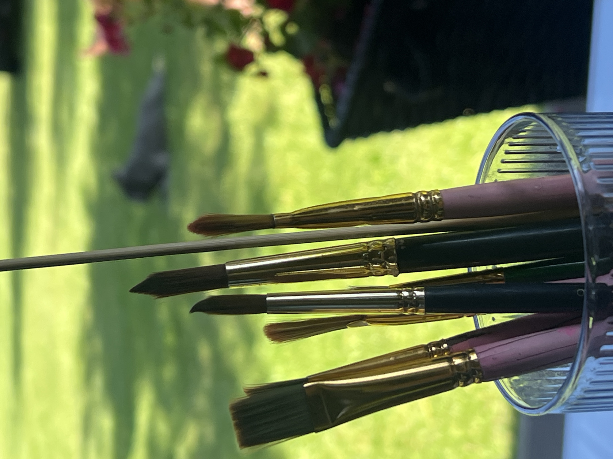 In the foreground of the image, a clear glass cup holds several paintbrushes. The cup has vertical ridges and is filled with the handles of the brushes, which are various colors including pink, black, and gold. The brush heads are also of different sizes and shapes. In the background, a green lawn is visible, with a blurred image of a grey cat lying down on the grass. To the right, a black planter with red flowers is partially visible. The overall scene appears to be outdoors on a sunny day.