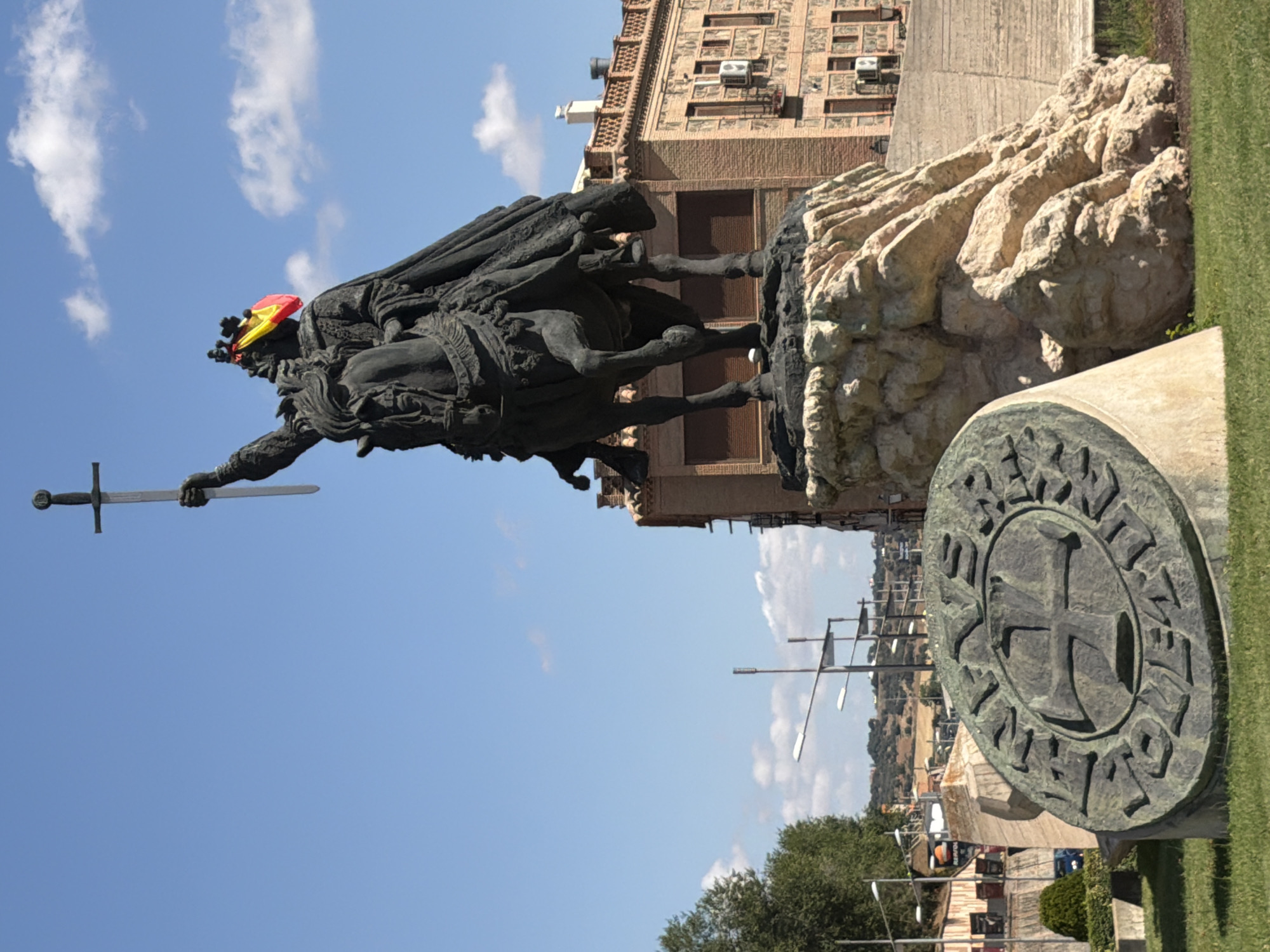 The image captures an equestrian statue of King Alfonso VI of León and Castile, prominently displayed in the city of Toledo, Spain, specifically in the Plaza deamoto. The bronze statue depicts the king on horseback, raising a sword aloft. A Spanish flag is draped over the king's crown, suggesting a patriotic or historical commemoration. The statue is mounted on a large rock formation, which is then set on a circular stone monument in the foreground. This monument features an inscription in what appears to be Old Spanish, with a prominent cross design at its center. In the background, a multi-story brick building with arched windows and balconies is visible, as are trees, streetlights, and a clear blue sky with scattered clouds. The overall scene suggests a historical monument in an urban or semi-urban setting.