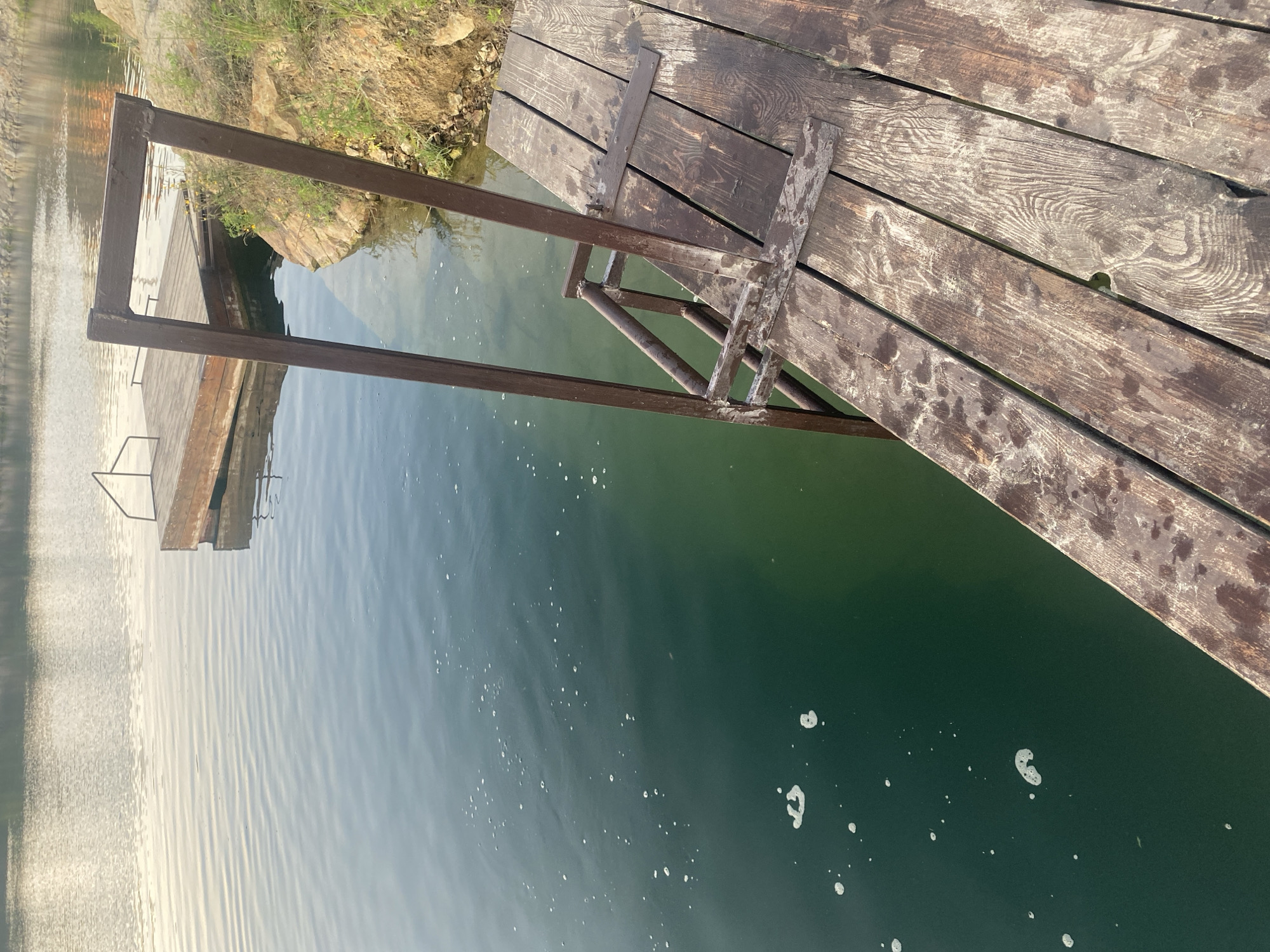 The image shows a rustic wooden pier extending into a calm, dark green body of water, likely a quarry lake, given the rocky embankment on the right. The pier features a dark metal ladder leading down to the water. In the background, another wooden platform is visible on the water. The scene appears to be a natural recreational area, possibly in or near Dnipro, Ukraine, judging by the style of the pier and the surrounding landscape which includes natural rock formations and sparse vegetation. There are no people or animals visible in the image. The lighting suggests it might be early morning or late evening, as the water reflects the soft light of the sky.