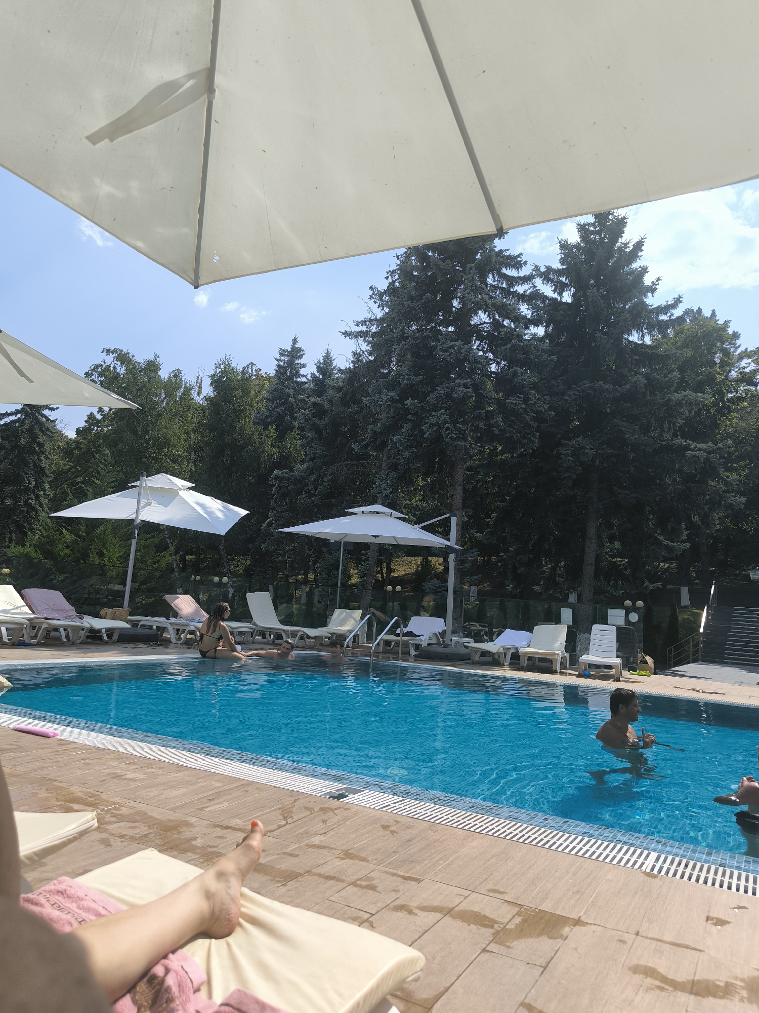 The image depicts an outdoor swimming pool area on a sunny day, likely at a resort or hotel in or near the city of Ungheni, Moldova, given the context provided by the user about Ungheni. The pool is filled with clear blue water. Several people are visible. In the pool, a man is standing waist-deep, holding what appears to be a drink with a straw. Another person is partially submerged nearby. Along the pool's edge, there are lounge chairs, some occupied by sunbathers. One person's leg and foot are prominently in the foreground, resting on a lounge chair with a pink towel. Several large white umbrellas provide shade over parts of the pool and seating area. The background features a dense line of evergreen trees, suggesting a natural or park-like setting surrounding the facility. The poolside is paved with light-colored tiles. Stairs leading up and out of the pool area are visible on the right side of the image.