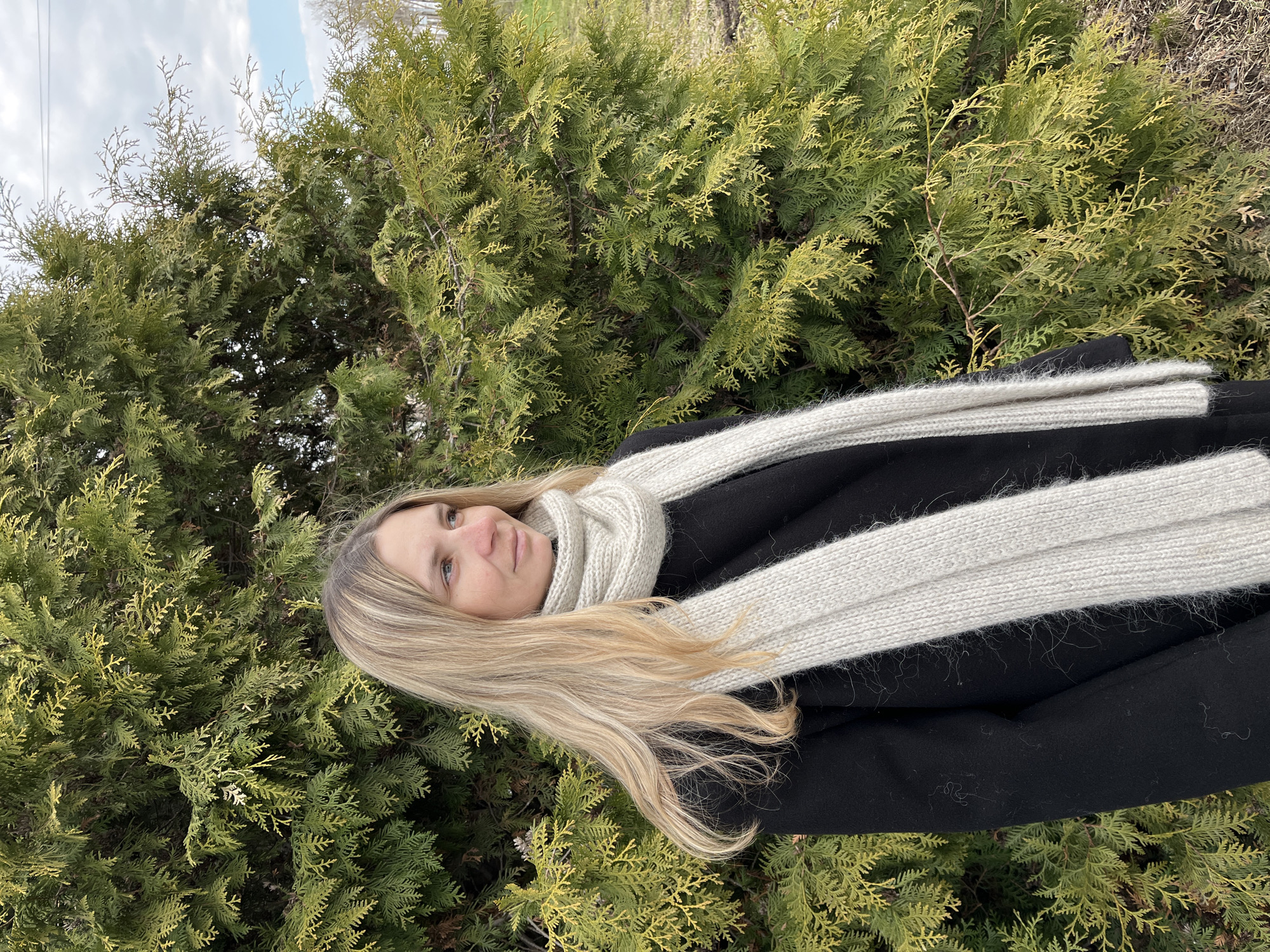 A woman with long, light-blonde hair, wearing a black coat and a thick, light-grey scarf, stands in front of a dense evergreen shrub. The sky is partially visible in the upper left corner, with patches of blue and white clouds. The location is Chaikovsky, Russia, based on available information.