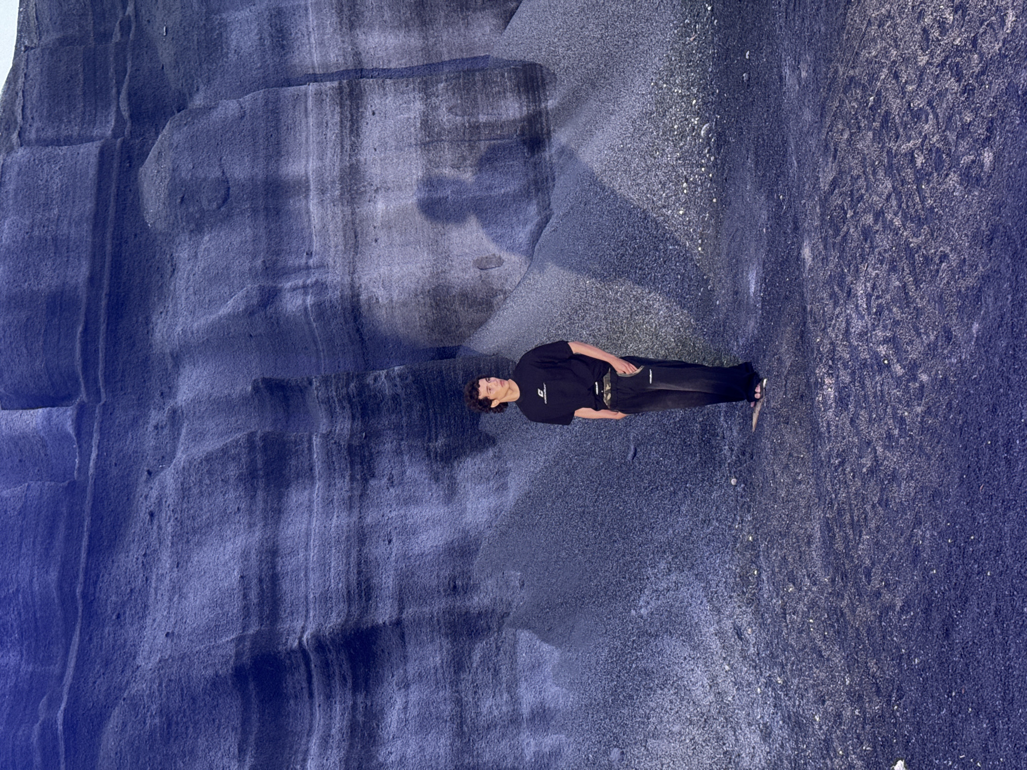 A lone individual, a young male with curly dark hair, stands within a stark, rugged landscape. He is facing the camera. The environment, characterized by its blue-gray tones, suggests a geological formation possibly made of volcanic rock and stone. The person is clad in a black T-shirt and matching black pants and his features are clearly visible. The background suggests a steep, rocky face or cliff formation, with textured surfaces. The overall perspective is slightly above eye level, presenting a wide angle view of the person within this geological setting. The setting is in Teguise, Spain.