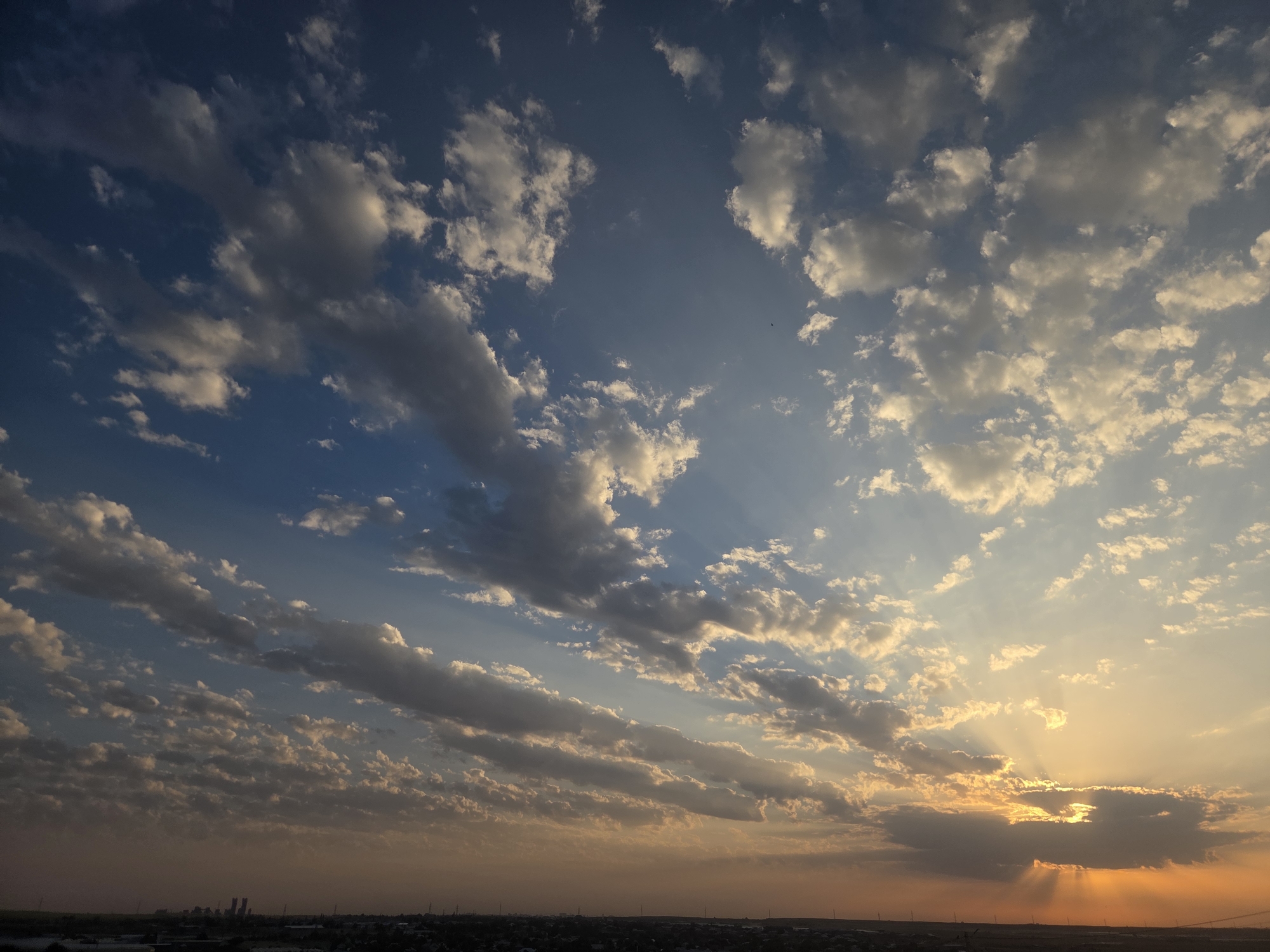 The image depicts a vast, dynamic sky over a cityscape, likely Қарасу, Kazakhstan, at either sunrise or sunset. The sky is a canvas of blues and oranges, with scattered clouds casting shadows and allowing rays of light to pierce through. The clouds vary in texture and density, from wispy streaks to heavier formations, some illuminated by the sun and others appearing darker. The sun itself is partially obscured by clouds on the right side of the frame, casting dramatic sunbeams downward. Below the sky, a distant horizon reveals a city silhouette characterized by low-lying buildings and structures, some of which appear to be industrial or institutional due to their scale and arrangement. The cityscape is muted in color, blending into the hazy atmosphere near the horizon. There are no visible people, animals, or specific actions taking place in the frame, focusing solely on the atmospheric conditions and the urban landscape.