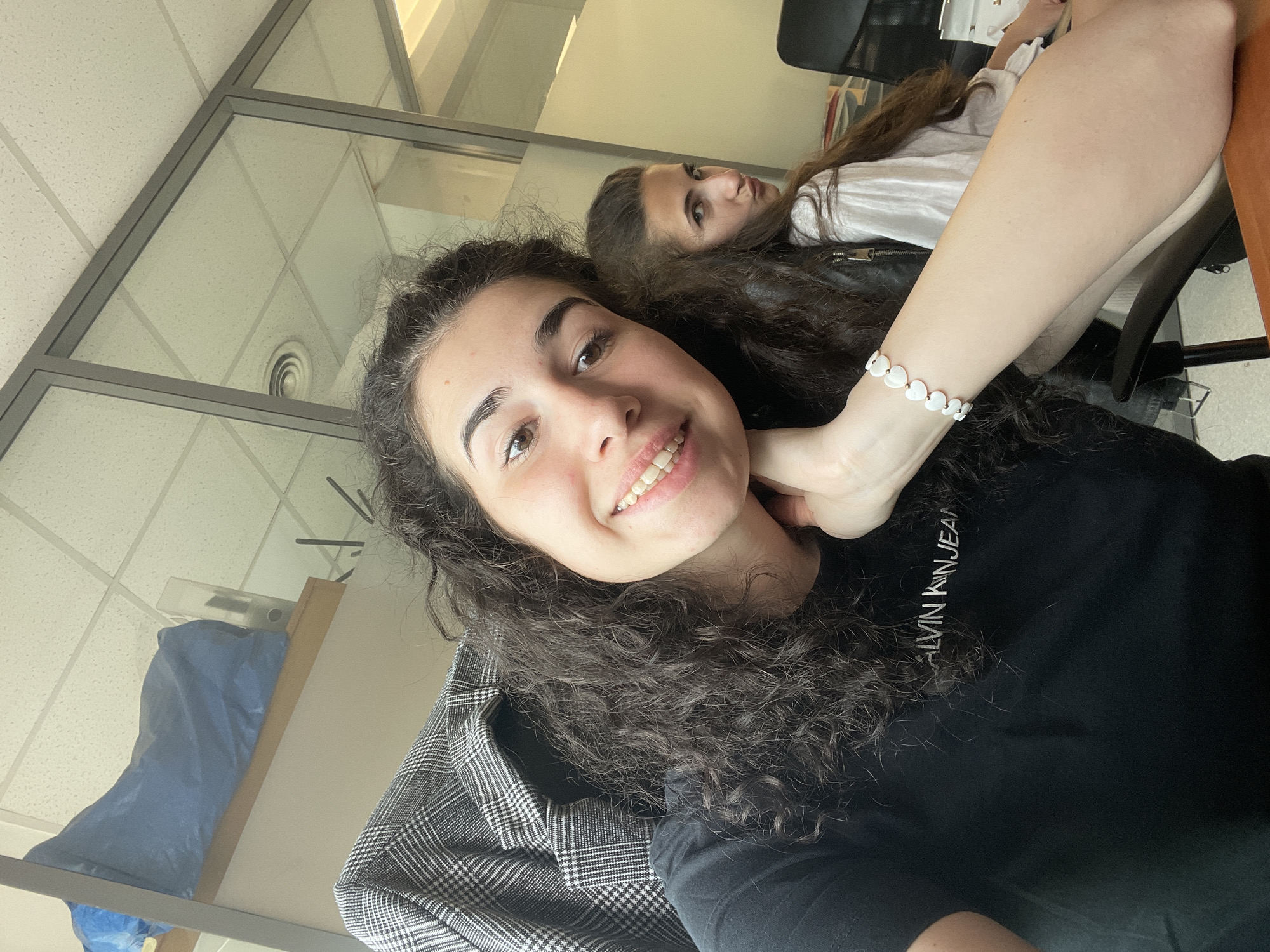 The image is a selfie of a woman with dark, curly hair, wearing a black Calvin Klein Jeans t-shirt and a white beaded bracelet. She is smiling. Another woman with long, dark hair is visible in the background, looking at the camera. The location appears to be an office space, evidenced by the ceiling tiles, office furniture, and glass partition in the background. There is no information present to determine that the location is in the city of Sofia, Bulgaria.
