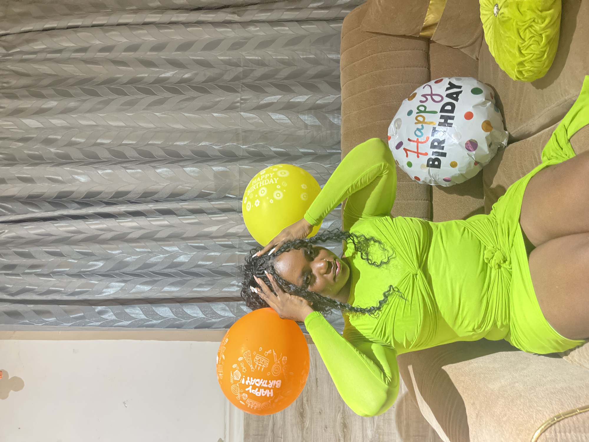 A young woman is captured in an indoor setting, likely in Kuchingoro, Nigeria, celebrating her birthday. She is lying on a brown upholstered couch, wearing a vibrant neon green, long-sleeved, ruched short dress. Her dark, braided hair frames her face, and her hands, with light-colored manicured nails, are resting on her head. She gazes towards the camera with a relaxed, gentle smile.
Three "Happy Birthday" balloons are prominent in the scene. One is a bright yellow balloon, another is orange, and the third is a white balloon featuring colorful polka dots. The text "Happy Birthday" is clearly visible on all three balloons; it's written in white on the yellow and orange balloons, which also include festive designs like cakes and party hats. On the white balloon, "Happy Birthday" is spelled out in multi-colored letters. A textured green cushion is also visible on the couch near her head.
The background consists of light grey curtains with a distinctive wavy or leaf pattern. Portions of a light-colored wall and what appears to be a floor or lower wall panel are also visible. The scene is brightly lit, suggesting it is daytime. The overall atmosphere conveys a cheerful and celebratory event.