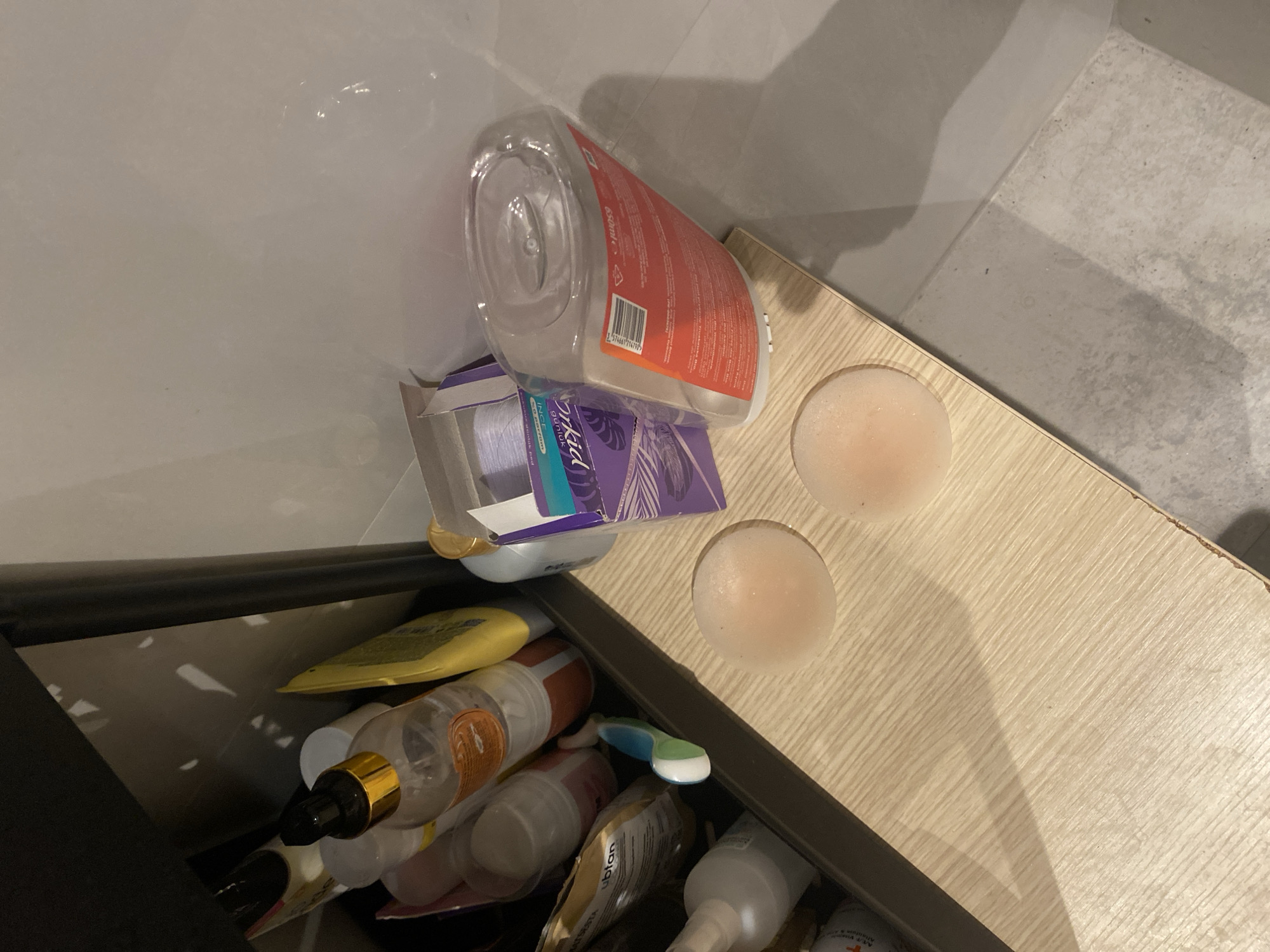 The image shows two light pink, circular silicone pasties on a light-colored wooden shelf. Behind the pasties, on the shelf, is a partially open purple box with white labels. To the left of the pasties, the shelf is filled with various bottles and containers, appearing to be bathroom or personal care products, including a clear bottle with an orange label and a black dispenser cap. The shelf is positioned in front of a dark shelving unit. The background consists of a light-colored tiled wall and floor. The image is an indoor shot, likely of a bathroom or vanity area. There is no indication of the city of Aksu, Turkey.