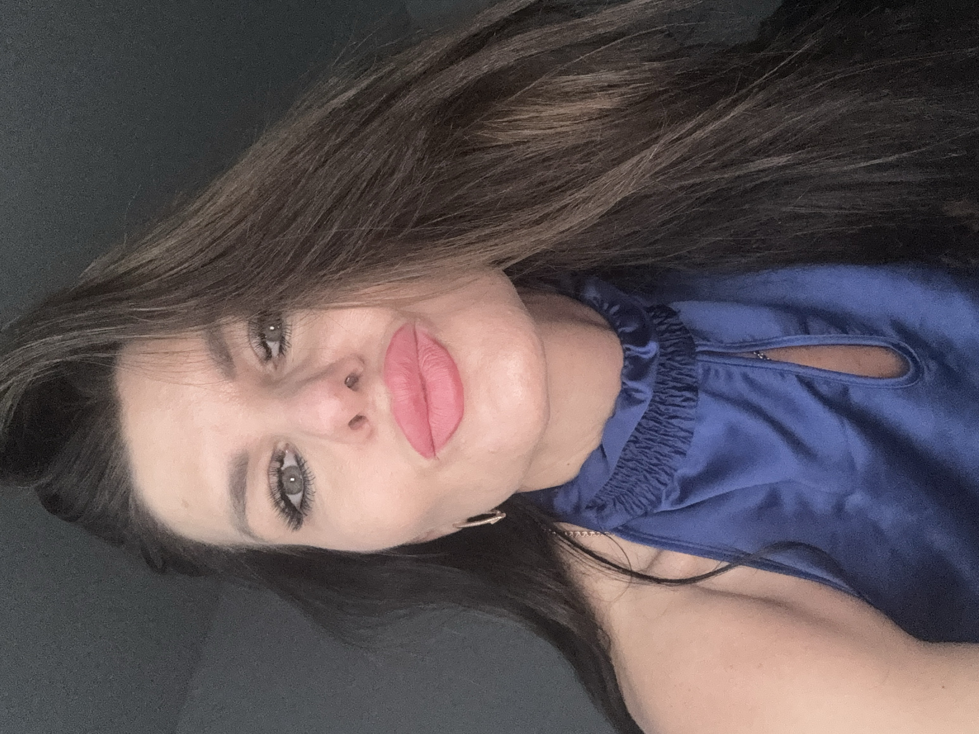 The media file is a close-up, vertically oriented photograph of a woman. She has long, dark brown hair that is partially obscuring her face and falling over her left shoulder. Her skin tone is fair. She is wearing a blue satin top with ruched detailing around the neckline and a small keyhole cutout. There appears to be a zipper visible within the cutout. She is also wearing a delicate necklace. A small hoop earring is visible on her left earlobe.
The woman's face is the primary focus. Her eyes are light-colored, and she is wearing dramatic eye makeup with prominent eyeliner and false eyelashes. Her lips are full and painted with a matte pink lipstick. Her expression is neutral, and she is looking directly at the camera.
The background is dark and appears to be a solid color, possibly a wall or furniture, with a subtle texture. The lighting is soft and appears to be artificial, suggesting an indoor setting. There are no clear indicators of the time of day, weather, or specific location beyond the general context of Chaikovsky, Russia. No text is visible in the image.