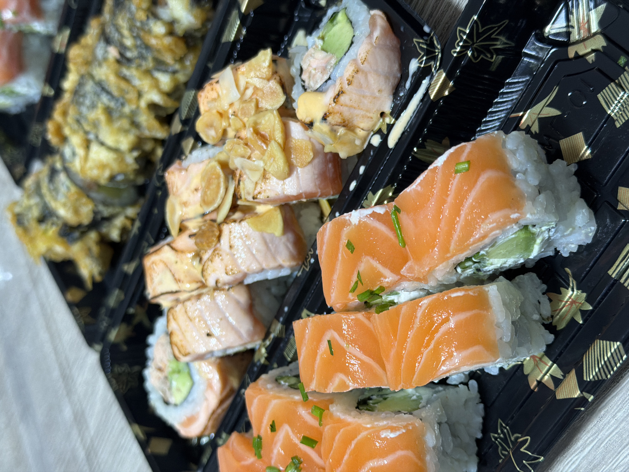 The image shows a close-up of three types of sushi rolls arranged in a black plastic container with a decorative gold maple leaf pattern, likely photographed indoors. The sushi appears to be in Ramat Gan, Israel, based on the metadata. The leftmost sushi is tempura-fried. The middle sushi is topped with seared salmon and fried garlic chips. The rightmost sushi is topped with raw salmon and a garnish of chopped chives. The rolls are filled with rice, avocado, and cream cheese.