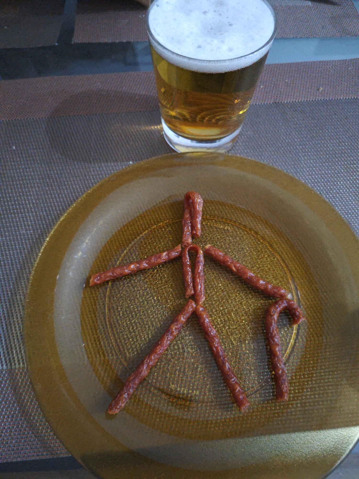 A glass of beer and a plate with a figure made of jerky sticks are on a table. The beer is in a clear glass with a foamy head. The jerky figure resembles a person, with a head made of two sticks, a torso made of one stick, arms made of two sticks, and legs made of two sticks. One leg is bent, and the other is straight. The figure is on a gold-colored plate, which is on a brown and grey placemat. The background is blurred and appears to be a dining room or kitchen.