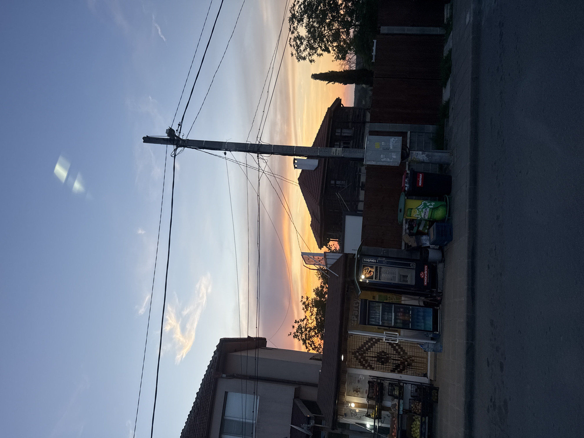 The image captures a street scene in Krushevets, Bulgaria, during sunset. The sky is painted with a gradient of soft oranges, pinks, and blues, indicating the end of the day. A tall utility pole with numerous wires dominates the left side of the frame, stretching upwards towards the colorful sky.
In the mid-ground, a traditional wooden house with a red-tiled roof stands against the sunset. Next to it, a modern building with shops is visible. This building has glass-fronted vending machines, one displaying the brand "lavim" and what appears to be a selection of drinks. Another machine has images of food items. A striped awning shades a storefront. There are also black trash bins situated near the vending machines. The street itself is paved, and a sidewalk is present in front of the shops.
To the right of the shops, a dark asphalt road is visible, suggesting a residential or commercial street. The overall atmosphere is peaceful, with the warm hues of the sunset creating a serene mood. The time of day is clearly sunset, and the weather appears clear, allowing for the vibrant sky. There are no people visible in the frame, focusing the scene on the architecture and the natural beauty of the sky.