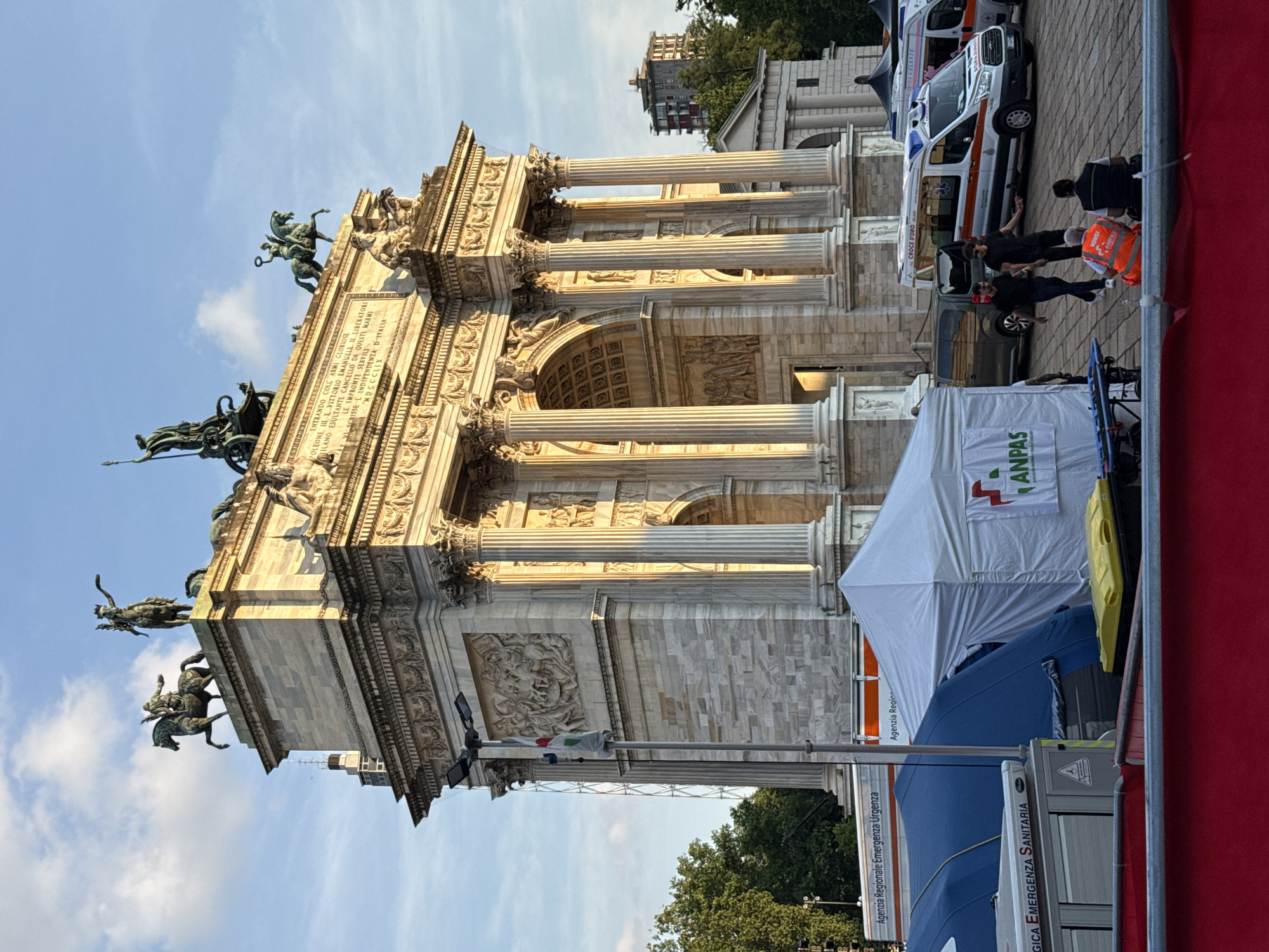 The image displays the Arco della Pace (Arch of Peace) in Milan, Italy, viewed from its eastern side, with a temporary emergency medical facility established in the foreground.
The Arco della Pace, a neoclassical triumphal arch constructed from light-colored stone, dominates the mid-ground. It features multiple arches, engaged columns with Corinthian capitals, intricate bas-reliefs, and various sculptures. The arch is crowned with a bronze Sestiga della Pace (chariot drawn by six horses) and other equestrian statues and figures. Inscriptions are visible on the arch's facade, including text mentioning "NAPOLEONE III" and "VITTORIO EMANUELE II." The arch is illuminated by direct, warm sunlight from the late afternoon or early evening, creating strong contrasts and shadows on its textured surfaces.
In the foreground, a temporary emergency medical setup occupies a paved plaza area. This includes a large white tent prominently displaying a red and green ANPAS (Associazione Nazionale Pubbliche Assistenze) logo, and at least two blue tents positioned behind it. A light gray trailer or truck with "Agenzia Regionale Emergenza Urgenza" printed on its side is visible behind the tents. Several emergency vehicles are parked to the right of the tents; one white ambulance with red and orange stripes and "CROCE D'ORO" is clearly identifiable, alongside others partially visible. A bright red barrier or structure runs across the extreme lower foreground. A yellow and black waste bin is situated near the white tent, and a stretcher is visible adjacent to it. An Italian flag (green, white, red) is unfurled on a tall pole positioned between the emergency setup and the arch.
Several individuals are present on the plaza. Two men in dark clothing are walking towards the right side of the frame, away from the tents. Another two individuals are seated on the ground in the mid-ground; one wears a high-visibility orange vest, while the other wears a dark vest over a dark shirt.
The sky above is clear blue with scattered white cumulus clouds. Green trees line the sides of the plaza and are visible behind the emergency setup, suggesting a park environment (Parco Sempione). Modern buildings are discernible in the distance behind the arch and trees.