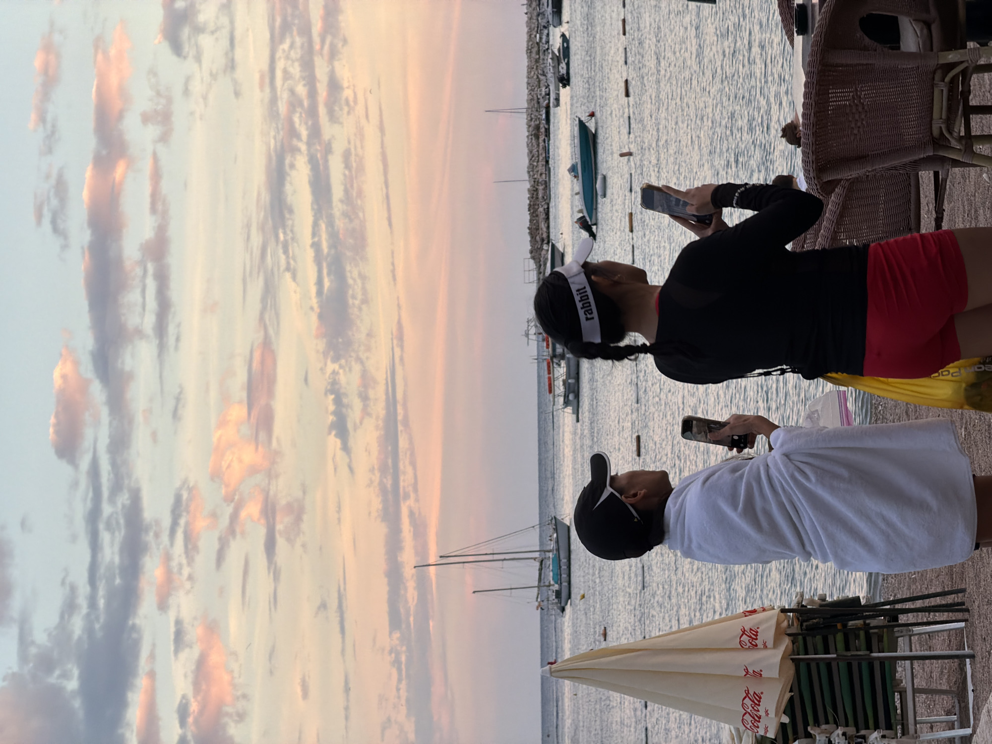 The image depicts a coastal scene at dusk in Budva, Montenegro. Two individuals are standing on a pebble beach, facing the water. The person on the left is wrapped in a white towel and is holding a smartphone, appearing to take a photo. They are wearing a black baseball cap. The person on the right, wearing a black long-sleeved top and red shorts, is also holding a smartphone and is looking out at the water. They are wearing a white visor with the word "rabbit" printed on it.
In the background, several sailboats and smaller boats are anchored in the calm sea. A breakwater made of large stones extends into the water on the right side of the frame. The sky is filled with soft clouds tinted with pink and orange hues from the setting sun. To the left, a cream-colored umbrella with a Coca-Cola logo is visible, along with beach chairs. The overall atmosphere is tranquil, suggesting a peaceful evening by the sea.