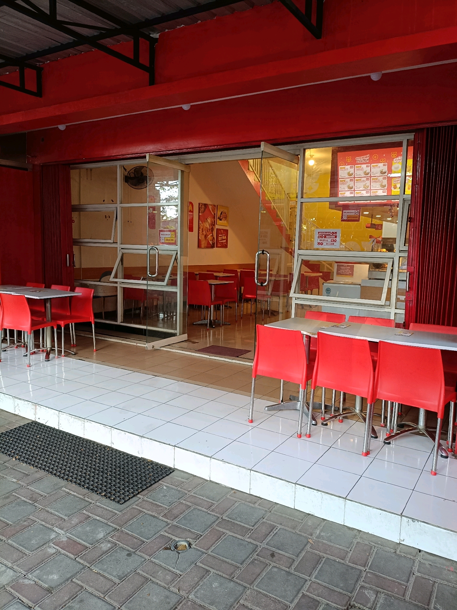 This is a medium-shot, eye-level photograph of the exterior of a brightly lit fast-food restaurant, likely located in Ngemplak, Indonesia, given the contextual clues in the image, such as the Indonesian text on signage and the architecture. The establishment is predominantly painted in vibrant red, with large glass windows and doors that offer a view into the interior.
The exterior features a red overhang supported by black metal beams. The facade is characterized by a glass storefront divided into sections. To the left, a set of tables with red chairs are arranged on a white tiled floor, creating an outdoor seating area. A black mat with a honeycomb pattern is placed at the edge of the tiled area.
Through the glass, the interior is visible, showing more red chairs and tables. A staircase with yellow railings is prominent in the background. Various promotional posters and menu boards with Indonesian text are displayed on the interior walls and behind the glass. One poster in particular displays food items and prices. Signs indicating "DILARANG MEROKOK" (No Smoking) are also visible.
The floor outside the restaurant is paved with interlocking stones in shades of gray and reddish-brown. A metal drain cover is visible on the paved area. The overall impression is that of a typical Indonesian fast-food eatery, likely busy during peak hours, but currently appears quiet with no customers visible in the outdoor seating area. The lighting suggests it might be daytime or early evening.