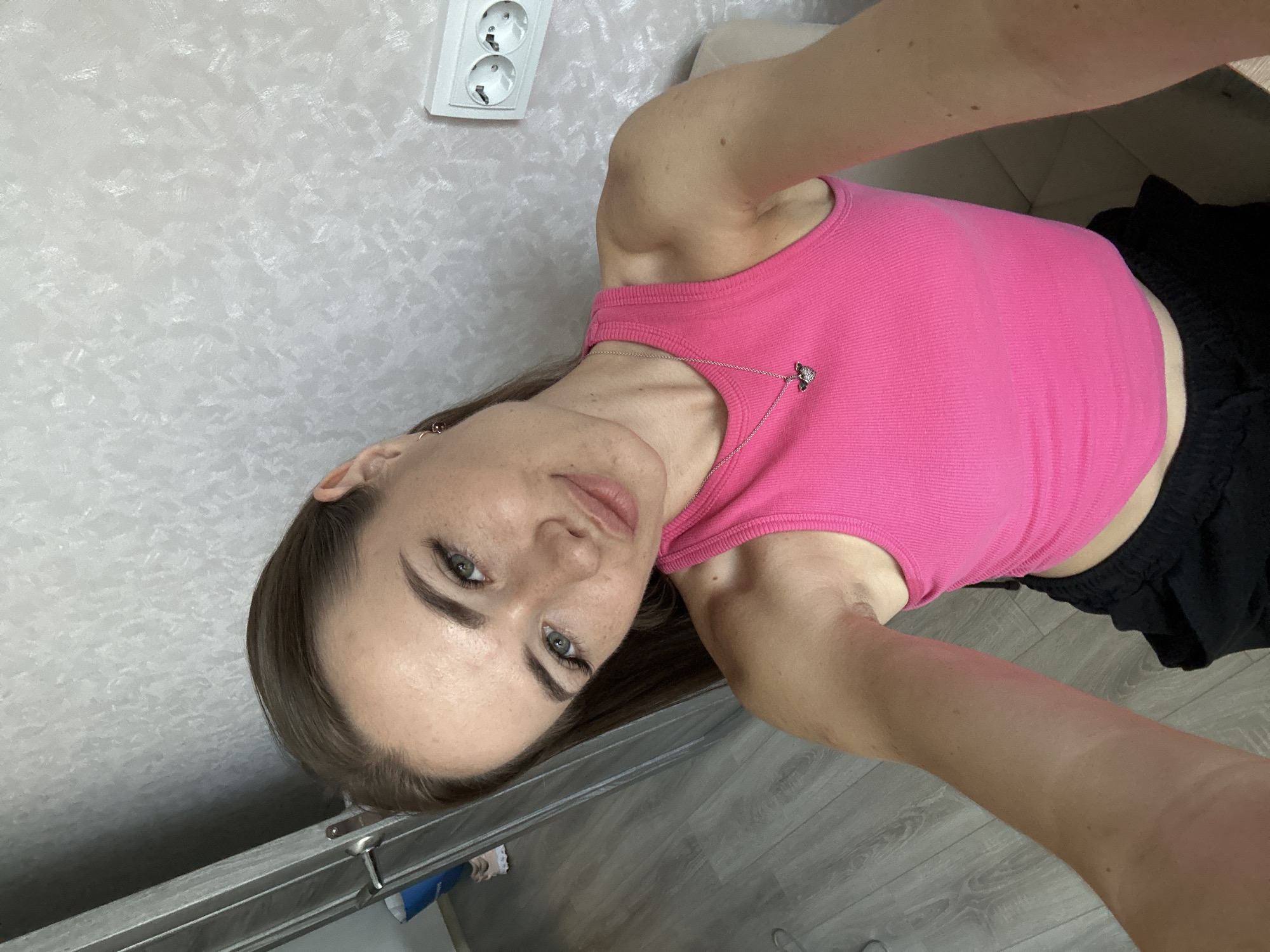 A young woman with light skin, brown hair, and green eyes is taking a selfie. She is wearing a pink tank top and black shorts. She has freckles on her face and arms. The background is a wall with a patterned texture and an electrical outlet. There is also a door and a wooden floor visible in the background. The woman is leaning on something or someone, and her left arm is extended towards the camera. Her expression is neutral, with her lips slightly pursed.