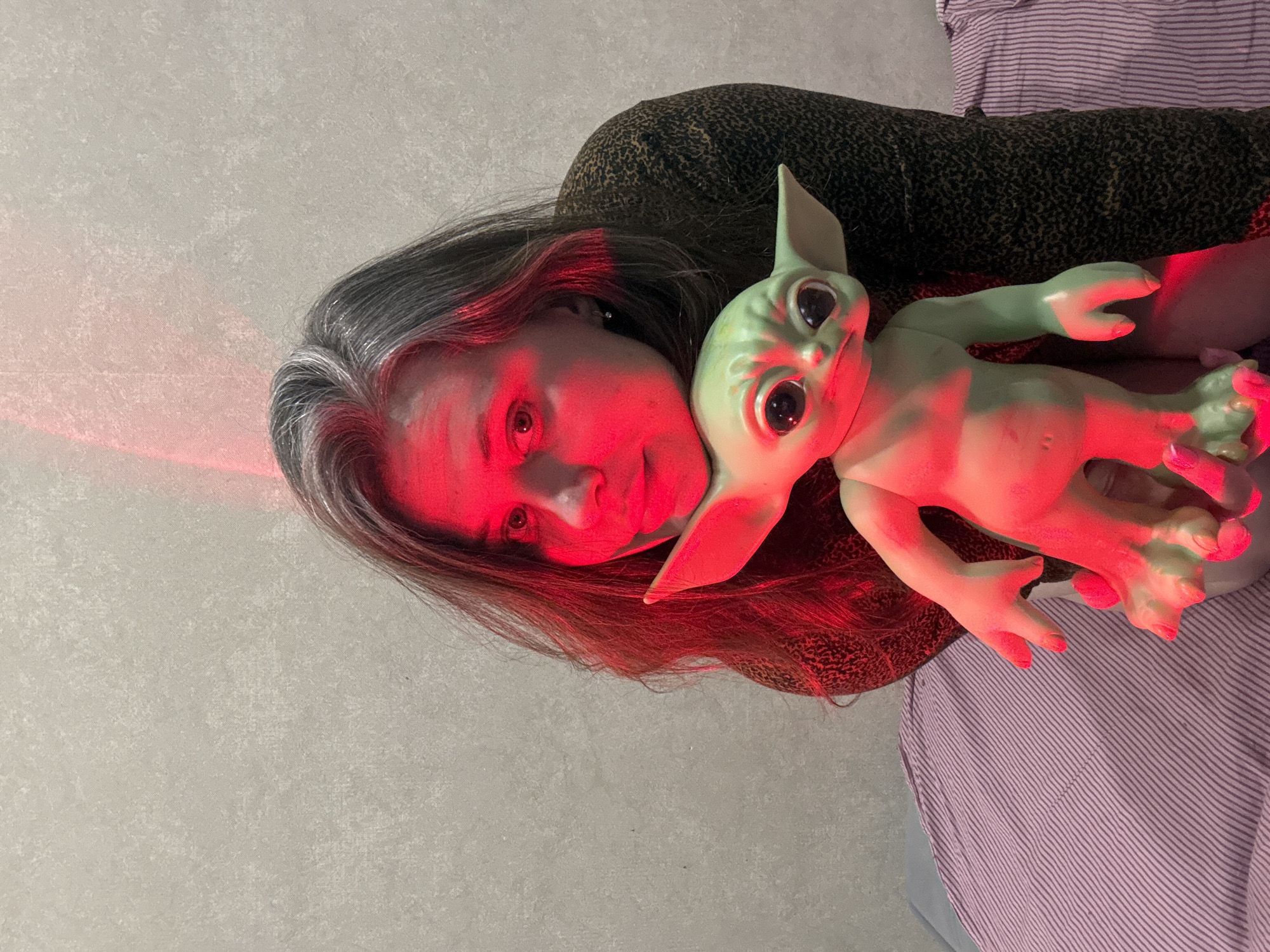 A woman with graying hair is holding a green, baby Yoda-like doll. Both are bathed in red light. The woman's head is tilted towards the doll. The background is a mix of a light-colored wall on the left and a striped fabric on the right. Part of the woman's dark animal-print clothing is also visible. Location is not discernible.