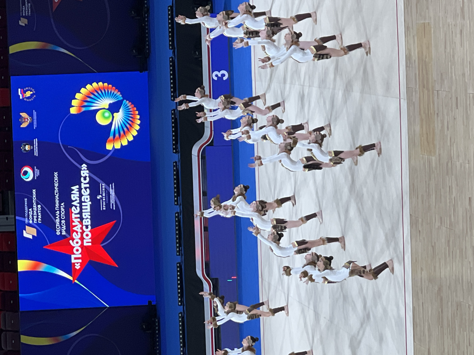 A group of gymnasts in white and brown uniforms are performing a routine on a white mat. The gymnasts are all in various poses of flexibility and balance, with many bending backwards. They have brown fur-like accents on their uniforms around their knees and waists. In the background, a large blue screen displays text and graphics related to an event, including a large red star with the words "ПОБЕДИТЕЛЯМ ПОСВЯЩАЕТСЯ" (Dedicated to the Winners). There are also logos and images on the screen, including a stylized peacock-like graphic. The floor in front of the mat is made of light-colored wood. A number "3" is visible on a blue section of the background behind the gymnasts. The lighting suggests an indoor venue with stage lighting.