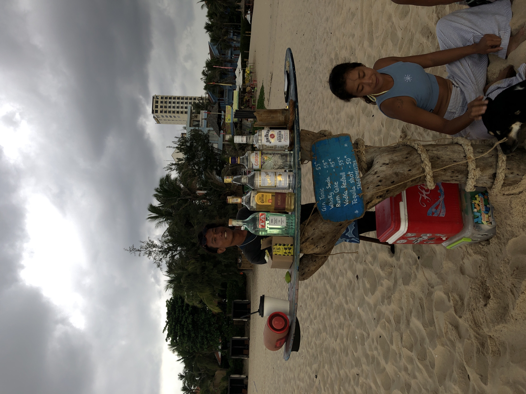 A man stands behind a makeshift bar on a sandy beach, displaying several bottles of alcohol, including gin, tequila, rum, and bourbon. A cooler, cups, and a speaker are also visible. A woman sits on the sand in front of the bar, petting a dog. In the background, palm trees, a hotel, and other beach structures are present. The sky is overcast. The location is Phu Thuy Ward, Vietnam.
