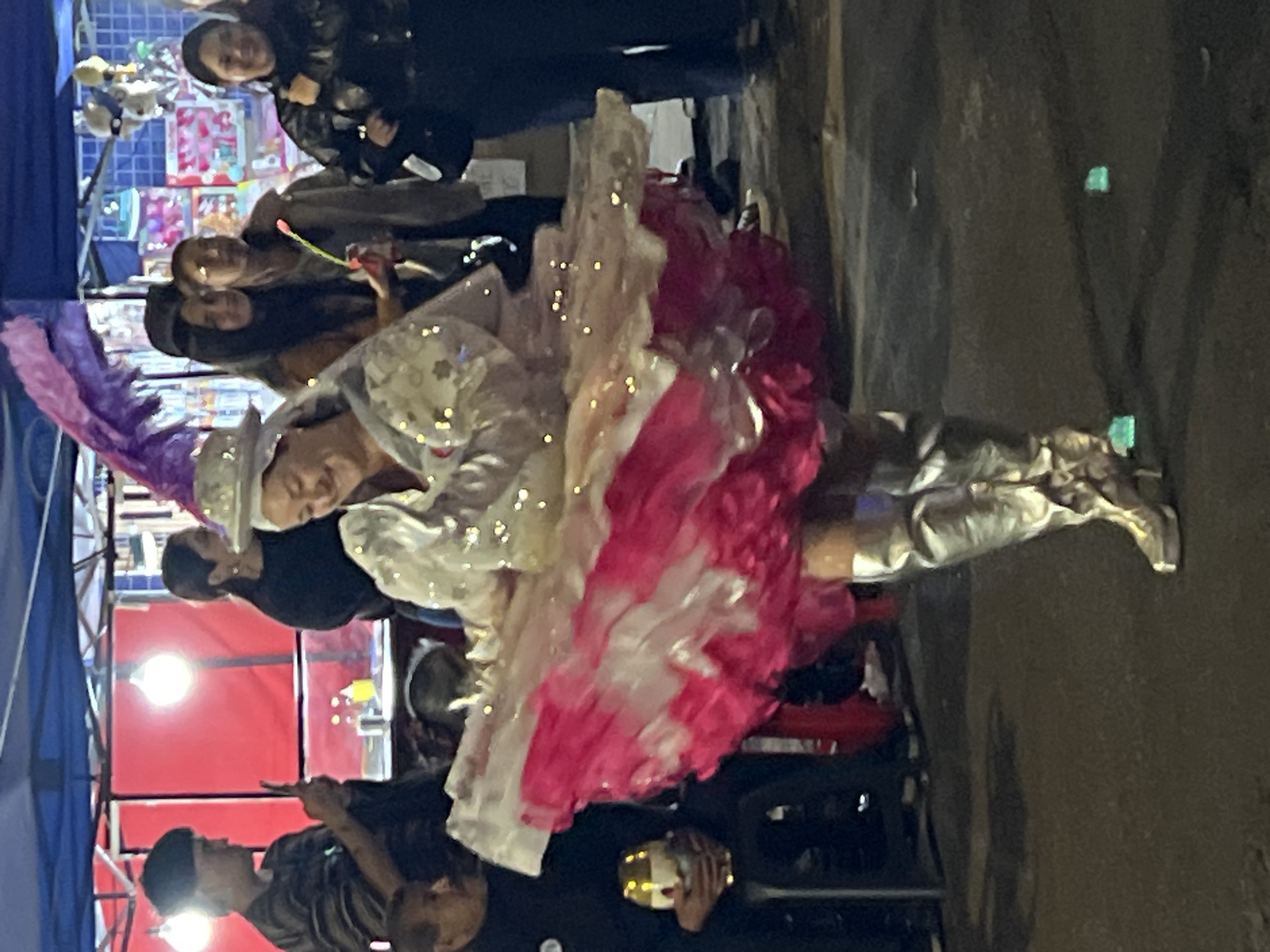 The image shows a woman in a shimmering white and pink costume, with a large, layered skirt and tall silver boots, in what appears to be a street performance or parade in Arica, Chile. She is smiling and looking down. She is wearing a white hat adorned with sequins and what appears to be a purple feather plume. Her costume is decorated with small, reflective embellishments that catch the light.
In the background, several other people are visible. To the left, a person in a dark top is holding a phone, possibly taking a picture. Behind them, a stall or tent with a red fabric backdrop is visible, illuminated by a bright light. Further back, other individuals are seen, some with blurred faces, suggesting a public gathering. The ground is paved and appears wet in some areas, reflecting the lights. The overall lighting suggests it is nighttime or dusk. The context of Arica, Chile, implies this scene may be part of a local festival or celebration.