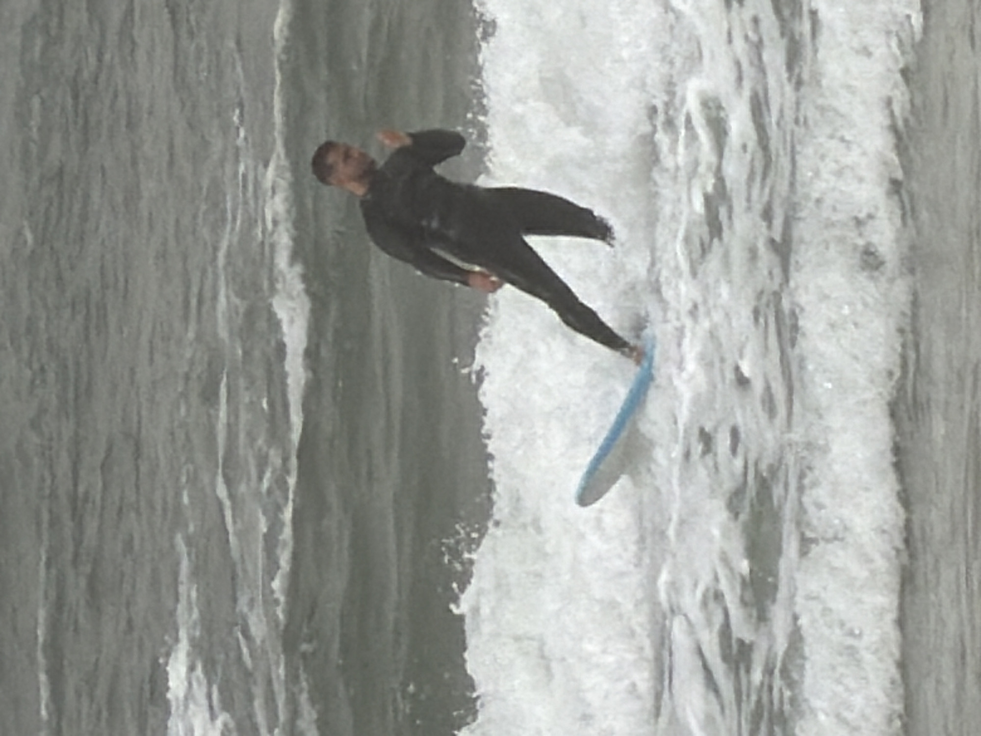 A man in a dark wetsuit is surfing on a wave. He is riding a light blue surfboard. The ocean is churning with white foam and gray water. The exact location in Tel-Aviv, Israel cannot be determined from the image alone.