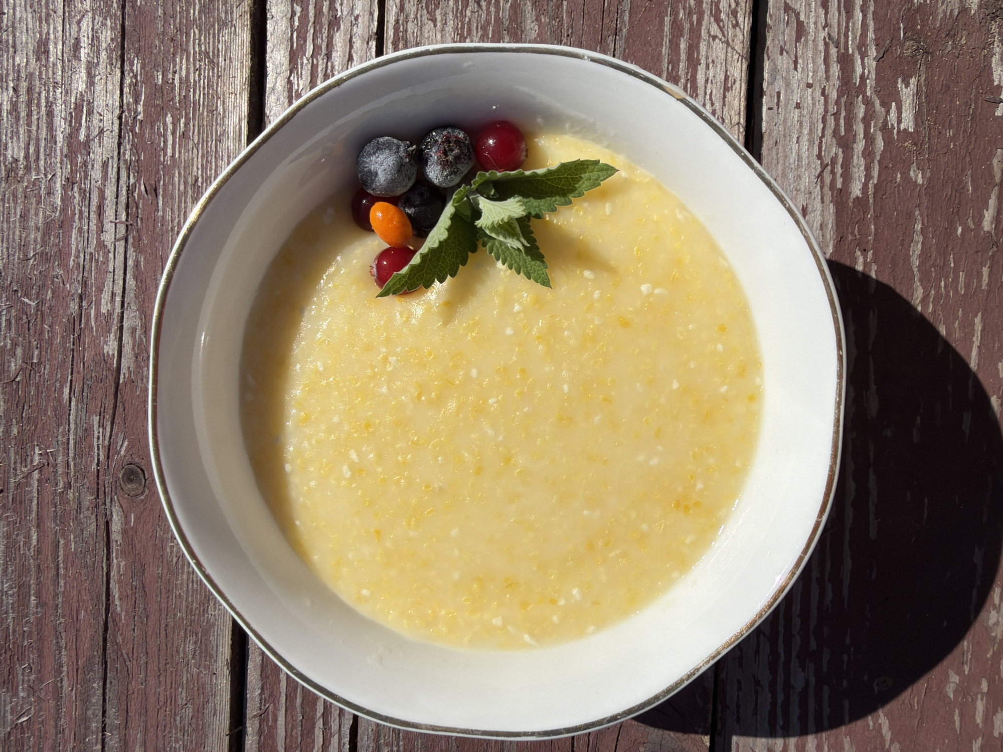 A white ceramic bowl with a gold rim sits on a weathered, dark red wooden surface. The bowl contains a pale yellow porridge, possibly cornmeal or semolina, with small white and yellow specks throughout. A small garnish of fresh mint leaves sits atop the porridge, accompanied by a few berries: two dark blue or black, frost-covered berries, two red berries, and one small orange berry. The wooden surface displays a distressed texture with visible grain and peeling paint. The scene is lit by bright sunlight, casting a dark shadow from the bowl onto the wooden surface. The location of Chaikovsky, Russia, is not discernible from the image itself.