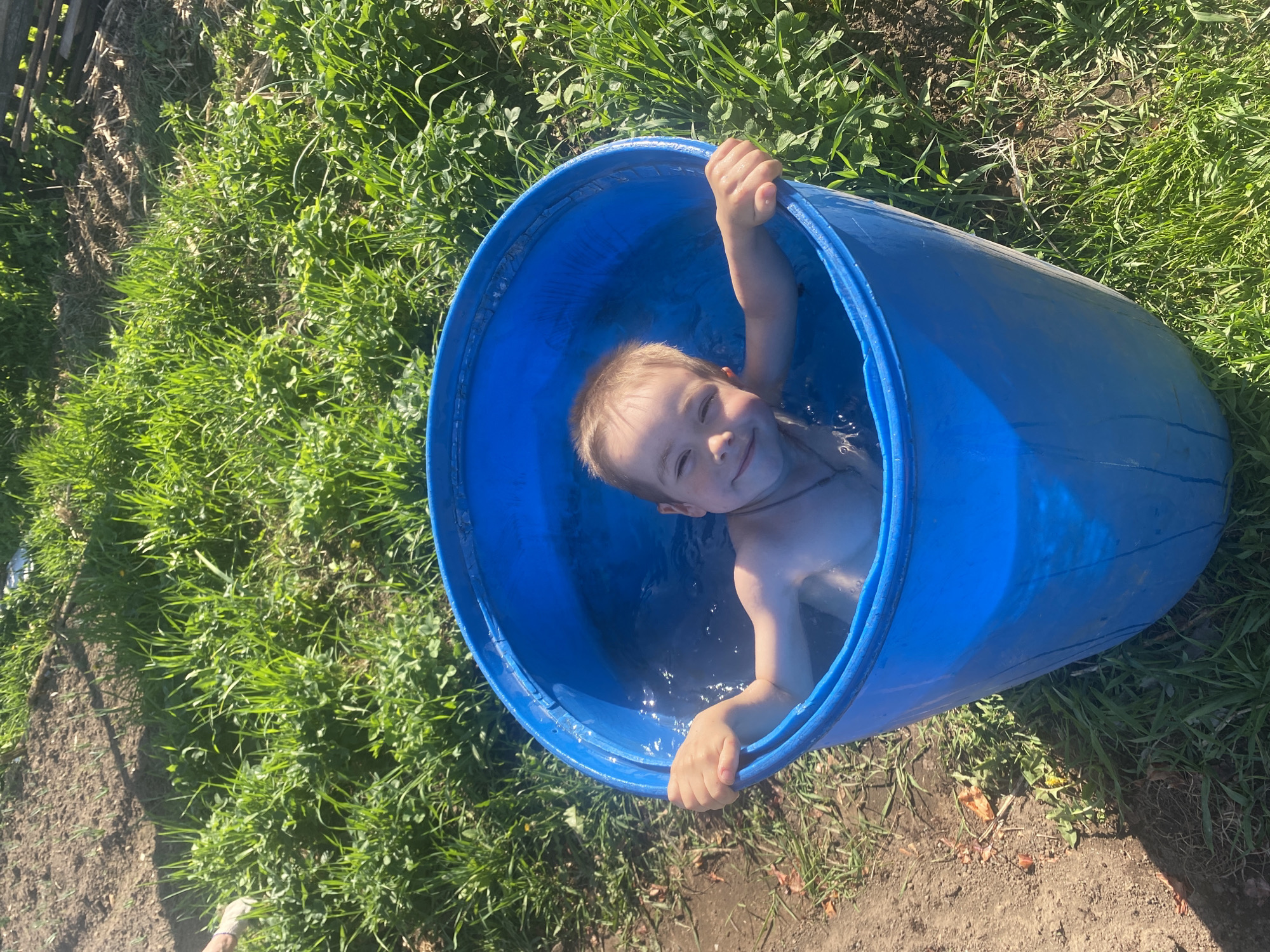 A young, fair-skinned child with short blonde hair is submerged in a blue plastic barrel filled with water. The child, appearing to be between the ages of 3 and 6, is positioned inside the barrel such that only their head, shoulders, and arms are visible above the water's surface. They are smiling directly at the camera and grasping the rim of the barrel with both hands.
The barrel is situated outdoors, adjacent to a patch of dense green grass. The ground surrounding the grass area is mostly bare and appears to be dirt or soil. The lighting is bright, suggesting the photograph was taken on a sunny day. Part of a wooden fence is visible in the upper left corner. Based on the visual content of the image, there is no verifiable information about the specific location of Узяр, Russia.