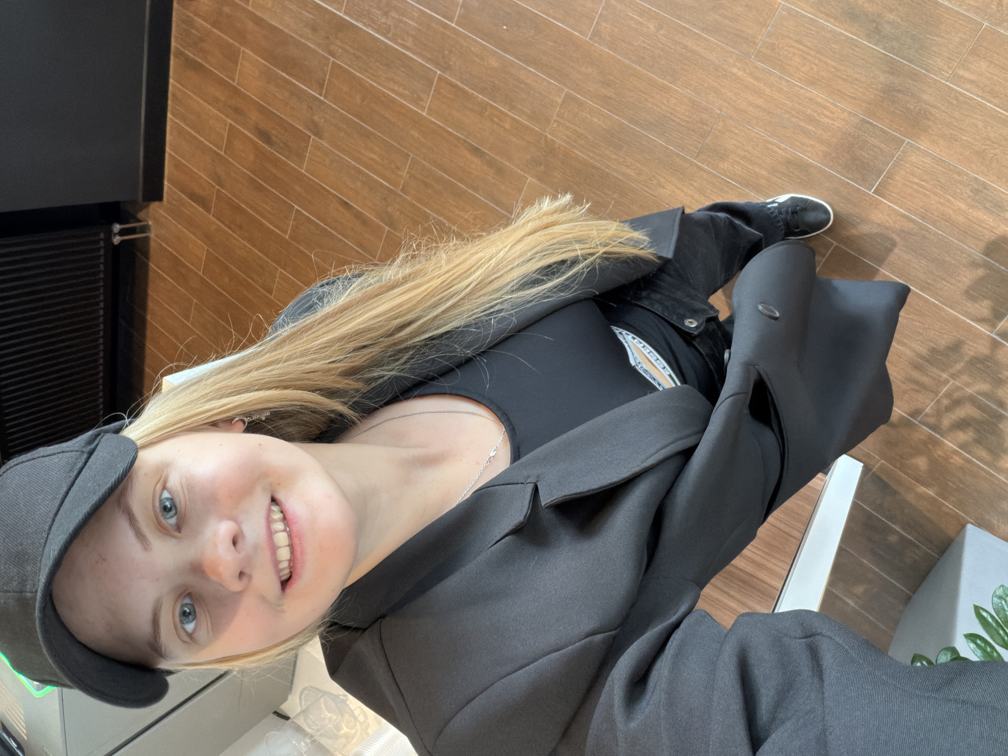 A fair-skinned young woman with blue eyes and long blonde hair is posing for a selfie. She is wearing a black cap, a black top, a jacket and jeans. She is seated on a chair and there is a section of wooden tile flooring. There are no discernible details to determine that the location is Izhevsk, Russia.
