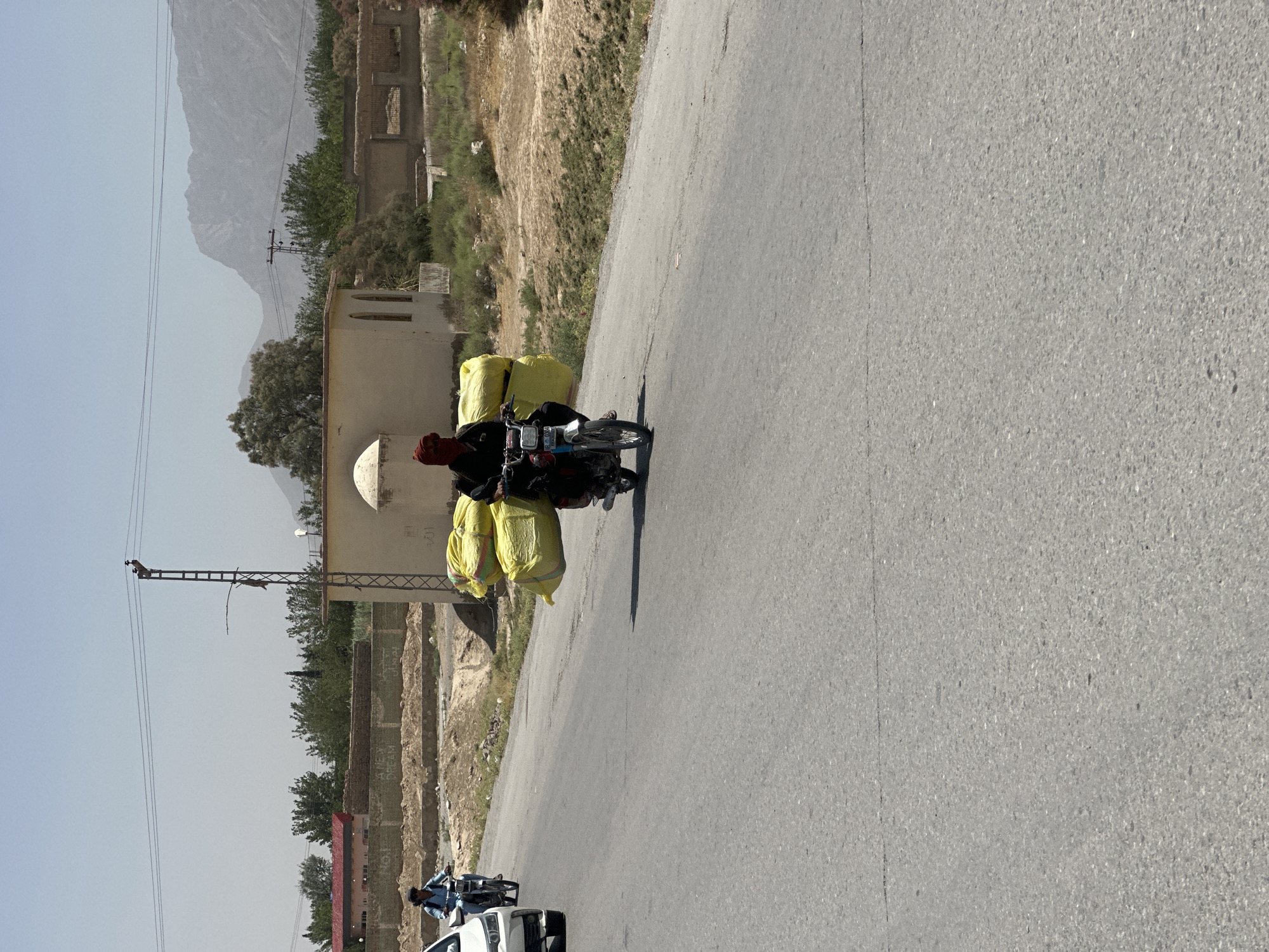 This media file captures a sunny, daytime scene on a paved road in a rural or semi-urban area, likely Kuchlak, Pakistan, given the context. The image features a person riding a motorcycle that is heavily laden with several large, bright yellow sacks, suggesting the transport of goods. The rider is wearing a dark outfit with a red head covering.
The motorcycle is positioned on the grey asphalt road, casting a visible shadow to its right. In the middle ground, on the right side of the road, the terrain is dry and covered with sparse vegetation. On the left side, there are simple buildings, including one with a distinctive small dome and some visible Arabic script on its wall, possibly "الله" (Allah). A tall utility pole with power lines is also prominent on this side.
In the background, majestic, hazy grey mountains dominate the horizon under a clear, bright blue sky, indicative of dry weather conditions. Further down the road, in the distance, another person on a motorcycle and a white car are partially visible, suggesting ongoing traffic. The overall impression is one of daily life and commerce in a mountainous, sun-drenched region.