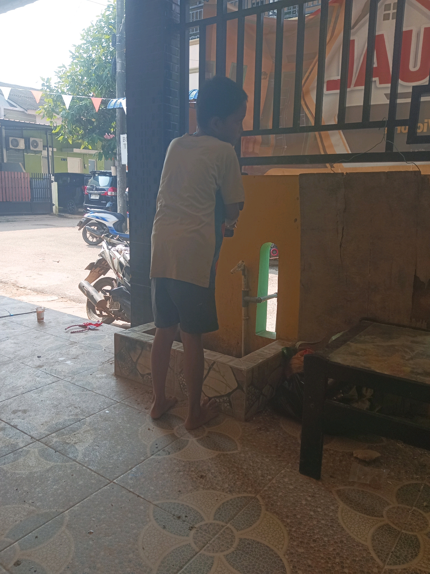 The image shows a young boy washing his hands at an outdoor faucet. He is wearing a light-colored t-shirt and dark shorts. The faucet is mounted on a yellow wall with a green, vertical opening behind it. In the background, there are motorcycles, a car, and houses. There are also festive flags strung between buildings. The scene appears to be in a residential area in Batam, Indonesia, likely a public space or a communal washing area. The ground is tiled, and there's a dark table-like structure to the right of the frame. The lighting suggests it is daytime.