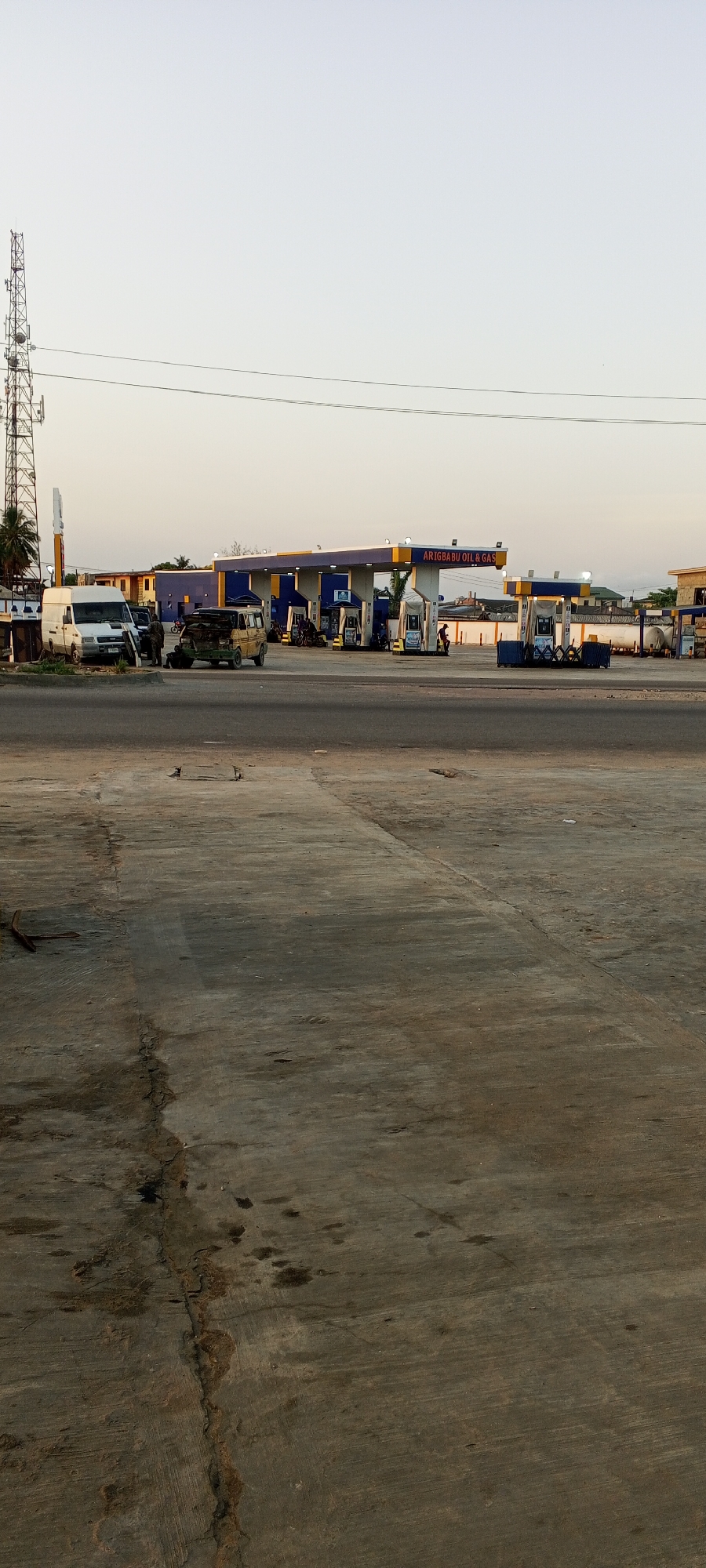 This media file depicts an active petroleum filling station identified as "ARIGBABU OIL & GAS" in Alasia, Nigeria. The scene is bathed in soft, diffused light, suggesting either early morning or late afternoon/early evening under a clear, pale sky.
In the foreground, a large, worn concrete or paved area dominates, showing numerous cracks and dark oil stains. The main subject, the gas station, is positioned in the mid-distance. It features distinct blue and yellow canopies sheltering multiple fuel pumps. Several vehicles are visible, including a white van parked on the far left and an older van with its hood open, indicating it might be undergoing repair or inspection, with an individual attending to it. Other vehicles are seen at the pumps, alongside several people who appear to be either refueling their vehicles or working at the station. A person on a motorcycle is also present near the pumps.
On the far left, a tall, metallic communications or cell tower stands prominently against the sky. Power lines stretch horizontally across the upper portion of the image. Behind the main gas station structures, storage tanks are visible on the right, and other buildings can be faintly discerned in the distance, providing a backdrop to the bustling but routine activity at the filling station.