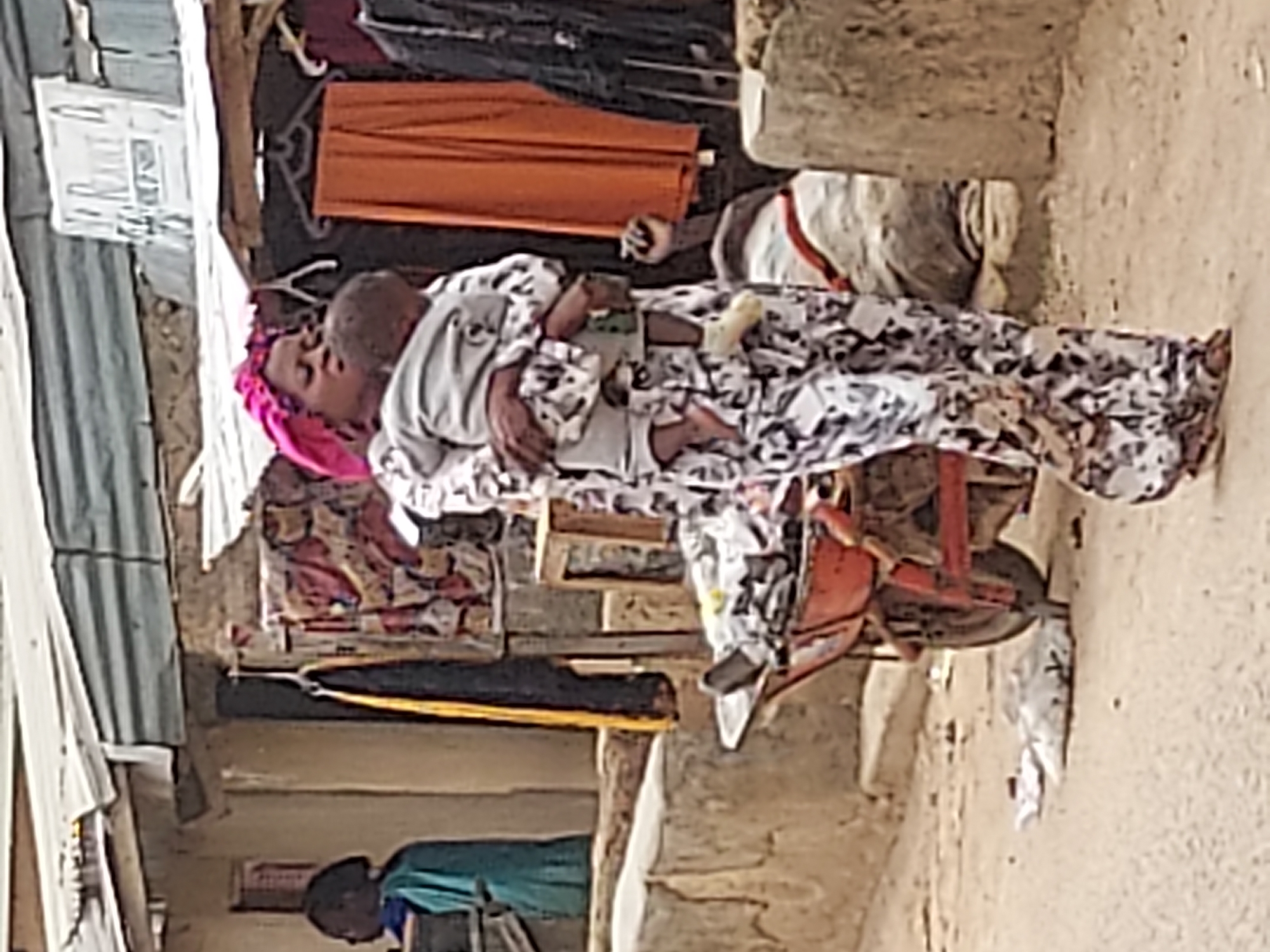 This image captures a quiet moment in Jos, Nigeria, featuring a woman holding a baby in an outdoor setting. The woman wears a vibrant pink headscarf and a long, patterned dress with black, white, and grey designs. She gently cradles the baby, who is dressed in a light-colored top and appears to be resting against her, looking away from the viewer. Her expression is calm.
The setting suggests a residential or market area with modest infrastructure. The ground is unpaved, appearing dusty and light brown. To the left, a structure made of corrugated metal sheets stands, on which a white sign with unreadable black text is affixed. In the background, an orange roll of fabric or material is prominently displayed, alongside various hanging clothes, hinting at commerce or domestic life. Another structure, possibly built from unrendered brick or concrete, forms the backdrop on the right. A reddish, three-wheeled object, resembling a small cart or wheelbarrow, is partially visible beneath the woman. Scattered small white fragments are strewn across the ground.
In the lower background, a third person is partially visible, wearing a green or blue top and looking downwards, seemingly engaged in an activity. The scene is bathed in bright, natural daylight, indicating it is daytime, likely mid-morning or afternoon, under clear weather conditions. The overall atmosphere is one of everyday life in a functional, community-oriented environment.