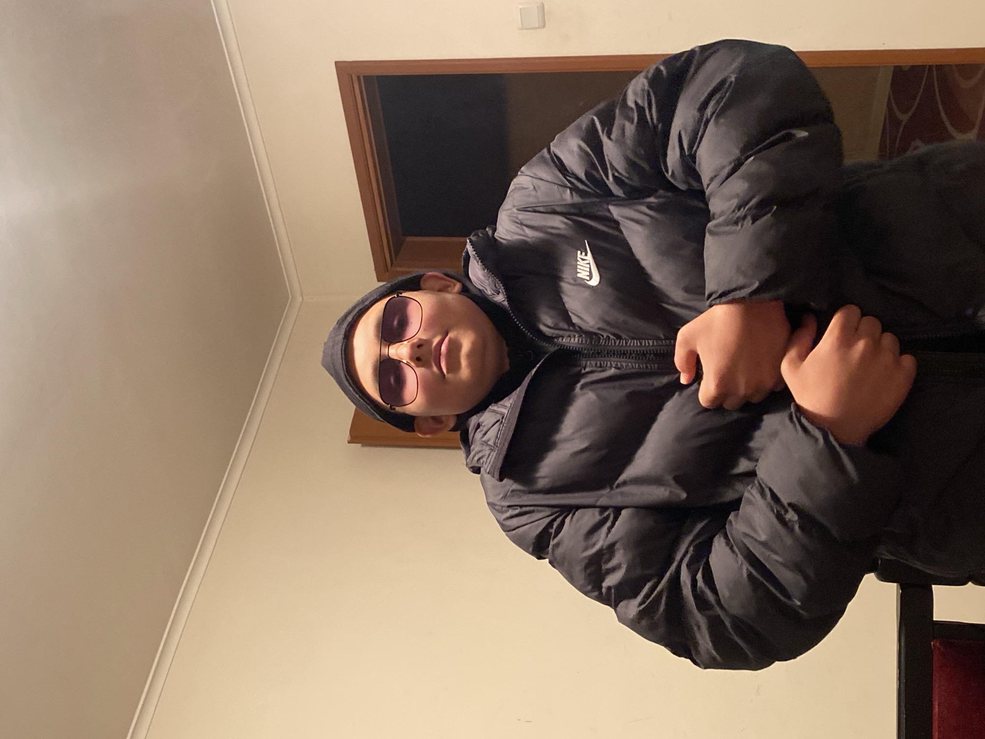 A person wearing a black beanie and rose-tinted sunglasses is positioned at an angle in the foreground. They are also wearing a black Nike puffer jacket. Behind them is a doorway with a brown frame. The background includes a plain, light-colored wall. The location cannot be determined from this image and there is no indication that it is in Goritsa, Bulgaria.