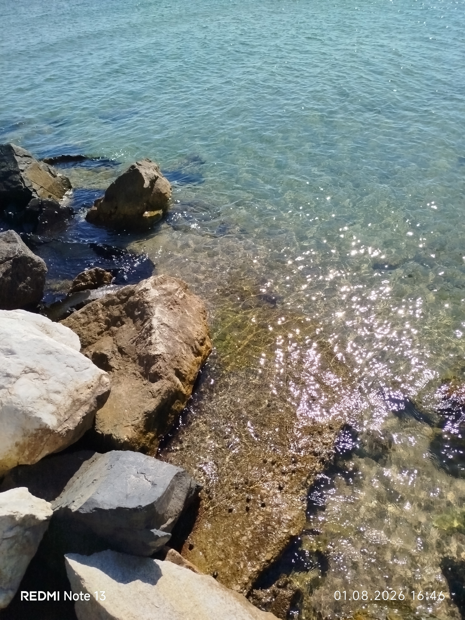 The image depicts a close-up view of the shoreline in the Sithonia Municipal Unit, Greece. The foreground is dominated by various rocks, some partially submerged in clear, shallow water. The water exhibits a turquoise hue and reflects sunlight, creating a shimmering effect on its surface. The rocks are of different sizes and textures, ranging from smooth, light-colored stones to rougher, darker ones. In the background, the open water extends to the horizon under a clear sky. The lower right corner of the image displays a timestamp, "01.08.2026 16:46," and the text "REDMI Note 13" is visible in the lower left corner, indicating the device used for capture. No people or animals are present in the scene.