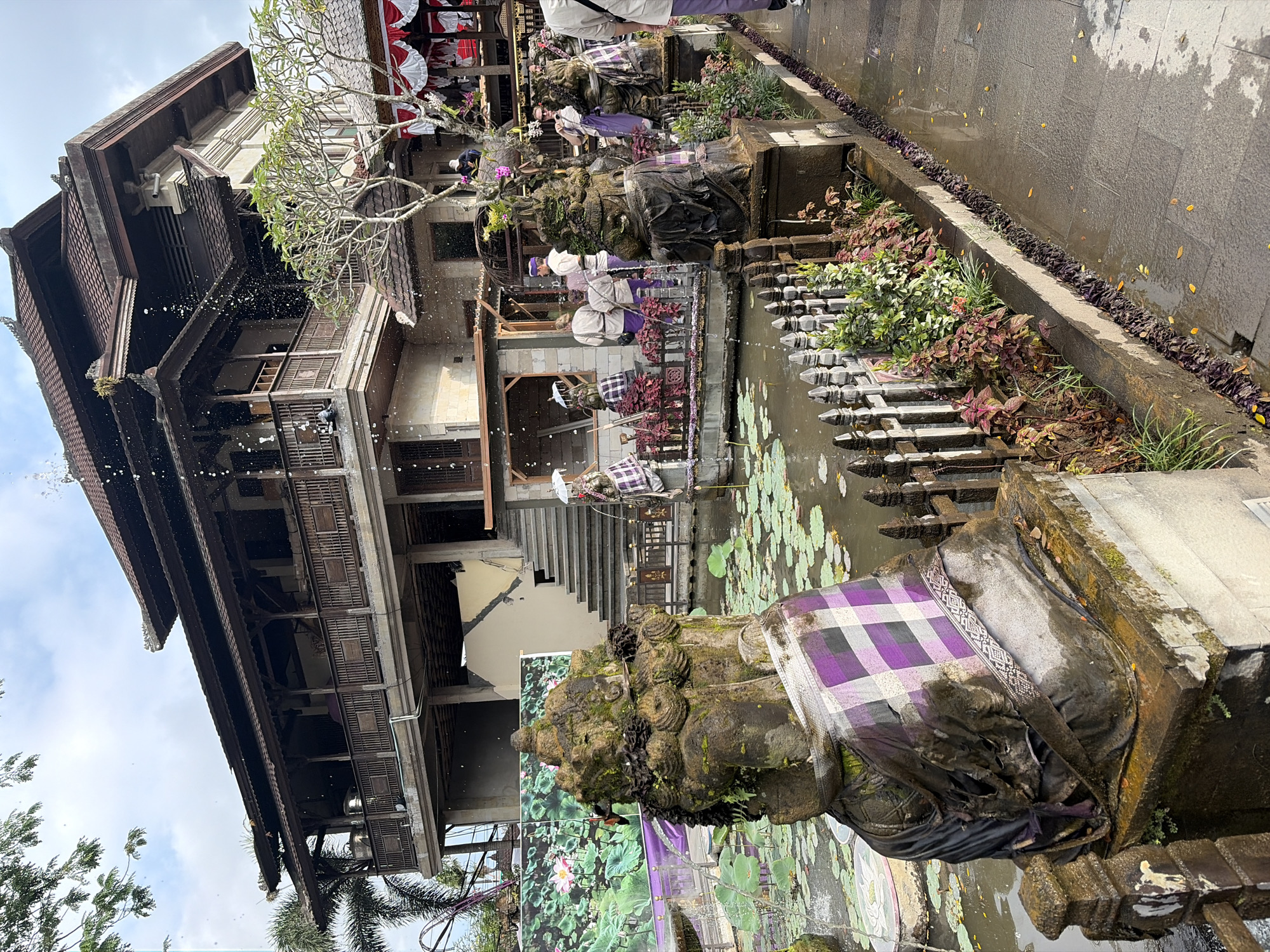 The image depicts a scene in Ubud District, Indonesia, characterized by a traditional Balinese building and a water feature. In the foreground, two moss-covered stone statues, adorned with purple and white checkered fabric, stand beside a lotus pond. The pond is populated with numerous lily pads and a few scattered flowers. Beyond the pond, a multi-story wooden and stone building with intricate carvings and a tiled roof dominates the background. Water is seen spraying from the building, creating a misty effect in the air. Several people are visible, some standing near the building, others in the vicinity of the pond, all appearing to be observing or participating in an event. The ground is wet and paved with stone tiles, suggesting it might have recently rained. The overall atmosphere suggests a cultural or ceremonial setting.