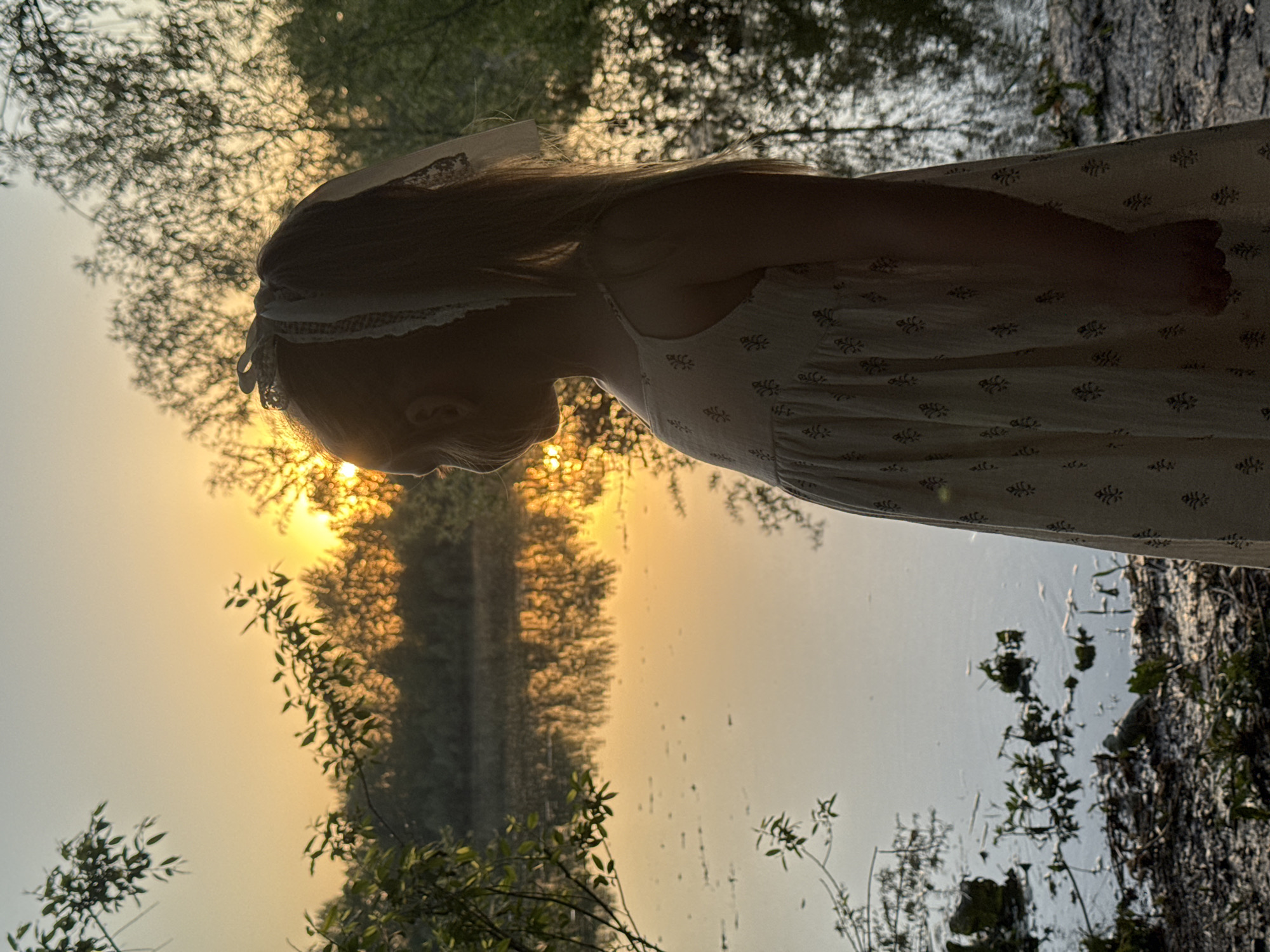 A young girl with long hair stands facing a body of water, possibly a lake or a wide river. She wears a light-colored dress with a subtle pattern and a headband. The sun is low in the sky, casting a bright golden light and creating reflections on the water. The environment is wooded, with trees lining the shoreline. The image is potentially taken near the city of Chaikovsky, Russia.