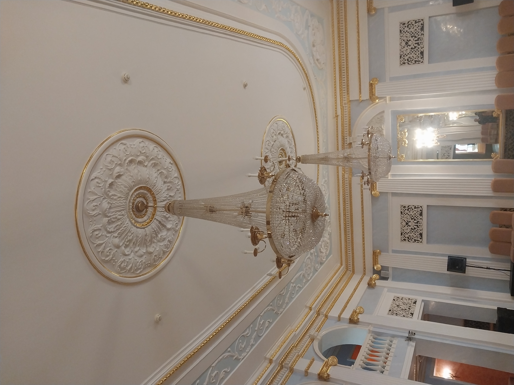 The image depicts the interior of a grand hall with ornate ceilings and elegant decor. Two large crystal chandeliers hang from the ceiling, casting a bright light. The ceiling itself is decorated with intricate plasterwork and gold trim, creating a lavish and luxurious atmosphere. Below the ceiling, the walls are adorned with decorative molding and panels, also featuring gold accents. In the foreground, the bottoms of several chairs are visible, suggesting that the hall is set up for an event or gathering. A large mirror on the right side of the image reflects the chandeliers and other elements of the room. The overall impression is one of opulence and historical significance. The location is identified as the Orenburg Regional Philharmonic Hall in Orenburg, Russia.