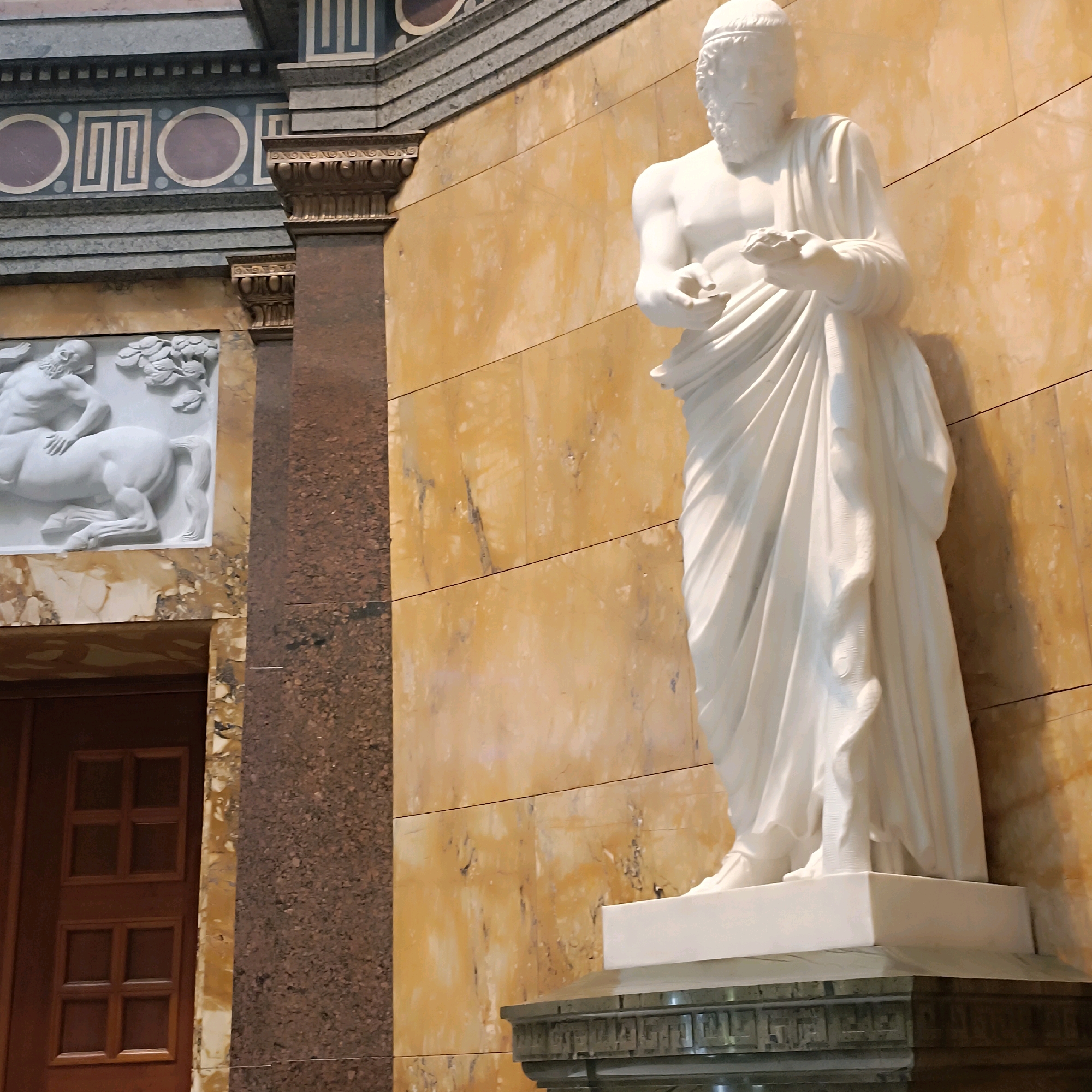 The image depicts a white marble statue of a robed male figure, likely an ancient Greek or Roman deity or philosopher, standing against a wall of veined yellow marble. The statue is detailed, with flowing drapery and a bearded face. A serpent is coiled around the figure's lower body, suggesting a connection to Asclepius or another mythological figure. To the left of the statue, a darker, textured pillar made of granite is visible, adorned with a carved frieze. Above the pillar, a decorative band with geometric patterns and circular inlays is set into a grey stone structure. Further left, a bas-relief sculpture of a centaur is partially visible. The overall impression is one of classical architecture and art. This statue is located in the Wiesbaden Casino, specifically in the foyer or a public area designed to evoke a sense of historical grandeur.