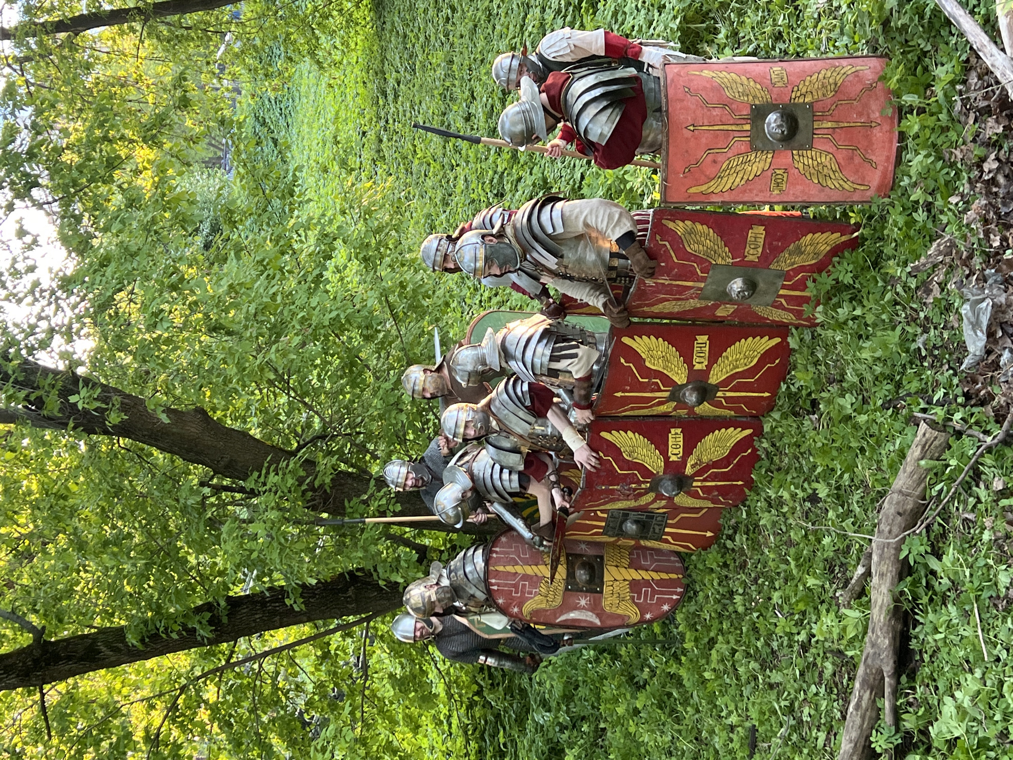 A group of people dressed in Roman legionary armor stands in a grassy, wooded area. The shields are red with a gold eagle design. The location is not identified.