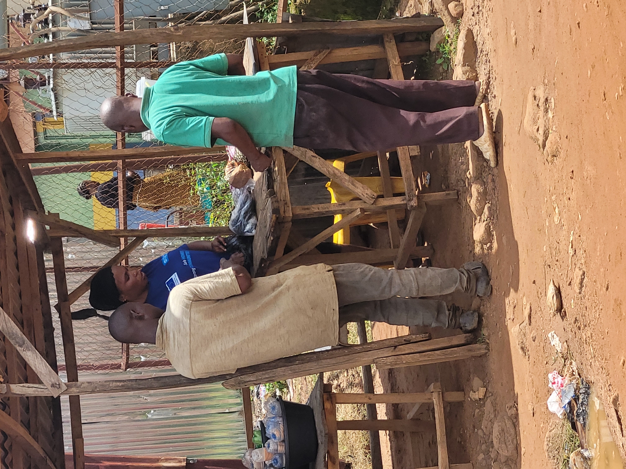 In Jos, Nigeria, under a makeshift wooden shelter, a woman wearing a blue t-shirt and a headscarf is engaged in a transaction with two men. One man, dressed in a light-colored t-shirt and gray pants, stands to her left, facing away from the camera. The other man, in a green t-shirt and dark pants, stands to her right, also facing away. They appear to be at an outdoor market stall. On a wooden table in front of them are various items, including what appear to be bags of goods and possibly some bottles. To the left of the stall, a black basin filled with bottled water is visible on a separate wooden table. The ground is dirt, with scattered rocks and some debris, including a puddle of muddy water with trash nearby. In the background, a chain-link fence encloses what looks like an electrical transformer and other industrial equipment. The overall scene depicts a street-side vendor in an informal commercial setting.