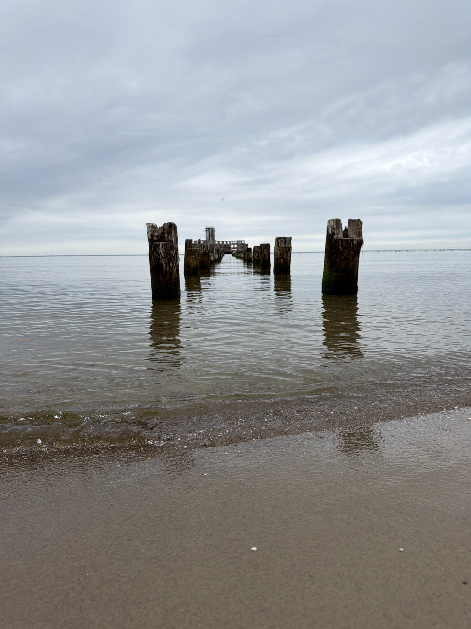 The image captures a desolate scene of an abandoned pier structure extending into a calm body of water. The pier consists of weathered wooden pilings, some of which are broken and moss-covered, leading towards a distant, dilapidated building that appears to be part of the pier's original construction. The water is still and reflective, with a gentle wave lapping at a sandy shore in the foreground. The sky is overcast with a thick layer of clouds, contributing to the somber and melancholic atmosphere. The location is identified as Unknown, Poland.