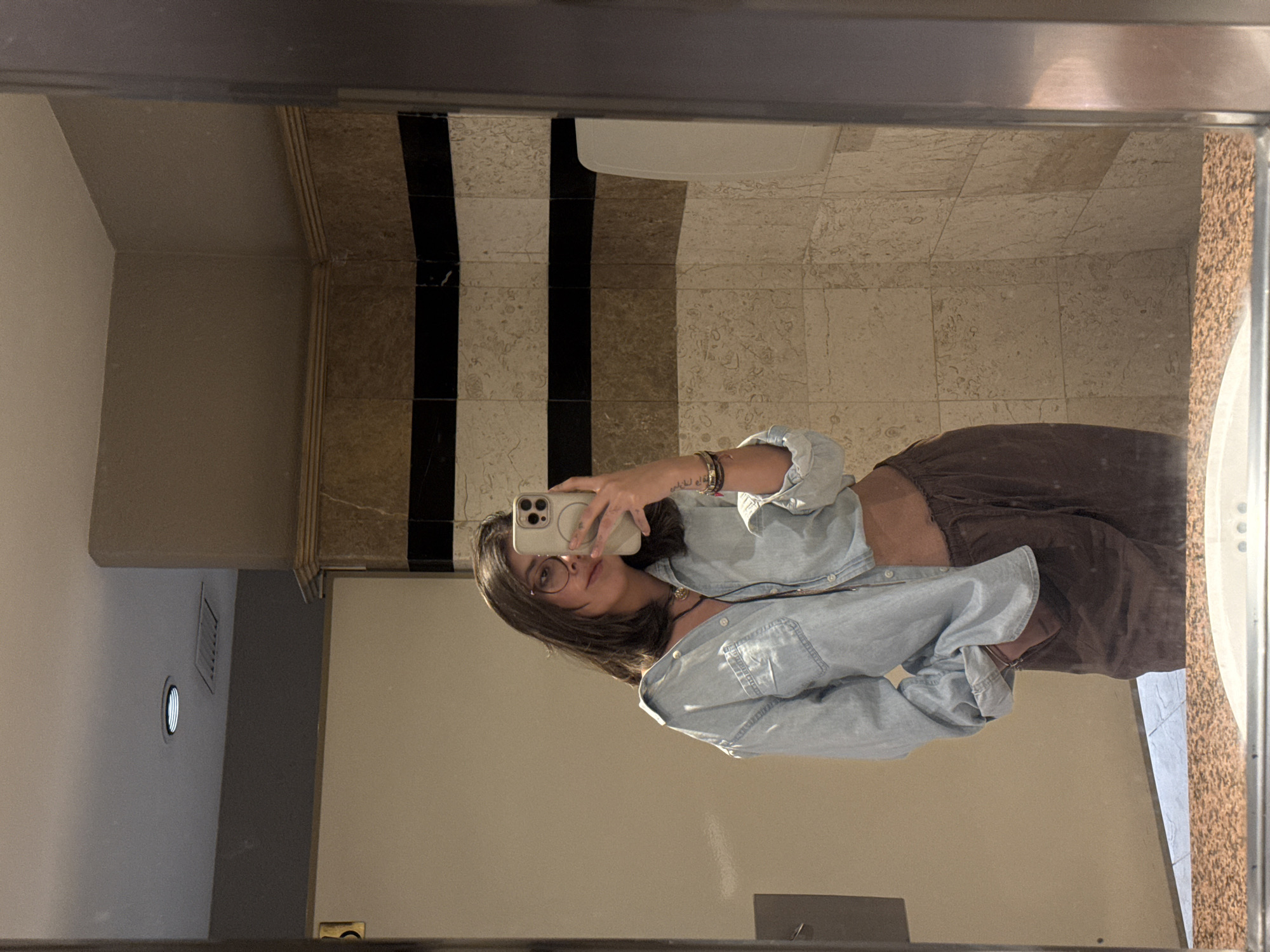 A female individual is captured in a mirror selfie within a public restroom in Puebla, Mexico. She is centered in the frame, wearing eyeglasses, a partially unbuttoned light blue denim shirt revealing her midriff, and dark brown loose-fitting trousers with her left hand in a pocket. A dark pendant necklace, multiple bracelets, and a tattoo with Arabic script are visible on her left forearm. She holds a light-colored smartphone with a multi-lens camera array to photograph her reflection.
The restroom features walls tiled with light-colored, possibly travertine or marble-patterned rectangular tiles, interrupted by two distinct horizontal bands of black tile at mid-height. Above the tiled section, a lighter beige painted wall is visible with a decorative molded trim. A white, wall-mounted dispenser or hand dryer is affixed to the right wall. The ceiling contains a circular recessed light fixture and a rectangular air vent. In the foreground, a speckled brown and orange granite-like countertop is visible, with the edge of a white sink basin. The floor, visible in reflection, is composed of light-colored square tiles. A light-colored door or partition is partially visible to the left. The overall environment suggests an upscale public facility.