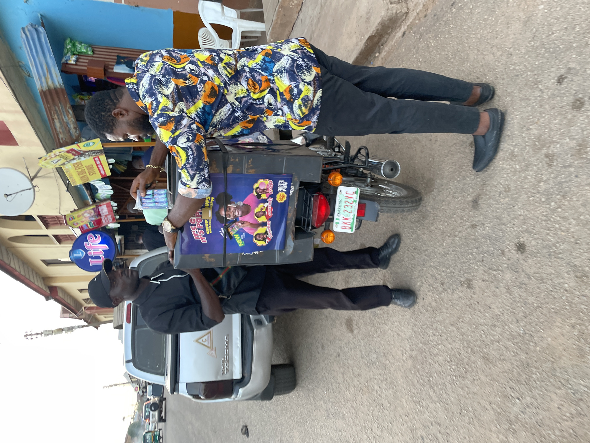 A street scene shows a man in a colorful patterned shirt standing beside a motorcycle, apparently stocking items on its rear cargo rack. The motorcycle has a license plate that reads "BKK-232VC". Adjacent to the motorcycle is a white Toyota Tacoma pickup truck, with a man wearing a black shirt and baseball cap standing by the truck bed. In the background, a storefront with a "Life" logo and advertisements for "Adon Big Fish Store" and "Chupa Chups" is visible. This scene is located in the city of Jos, Nigeria.